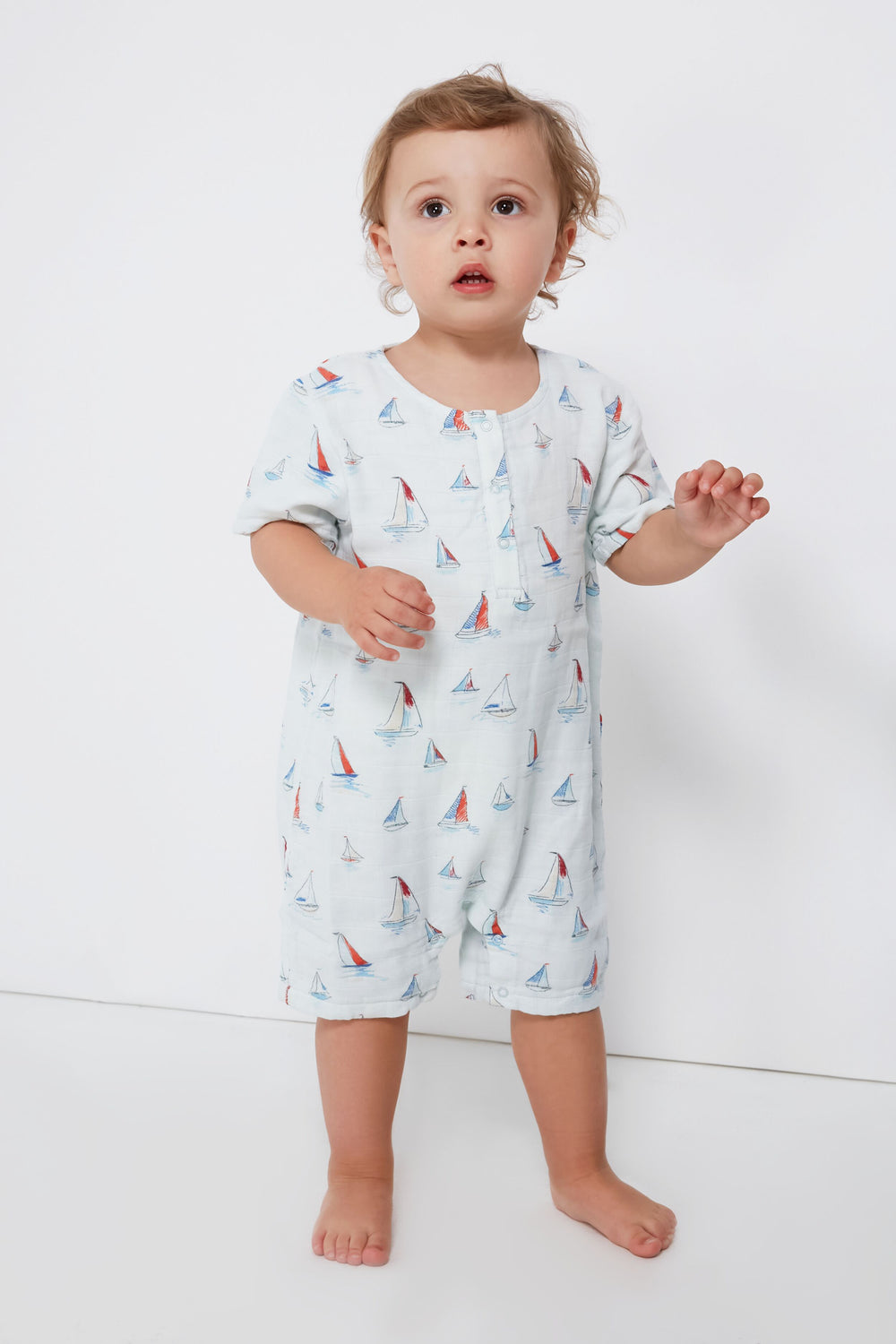 A toddler stands barefoot, wearing a light-colored organic muslin sailboat-print romper from the Angel Dear baby clothing collection.