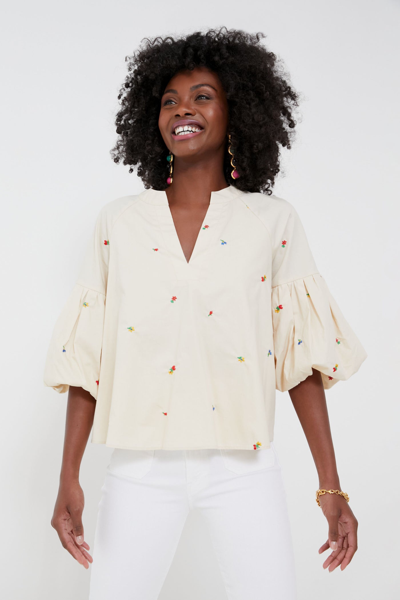 A person wearing a cream-colored, puff-sleeve embroidered blouse with colorful details from Hyacinth House, paired with white pants, stands smiling.