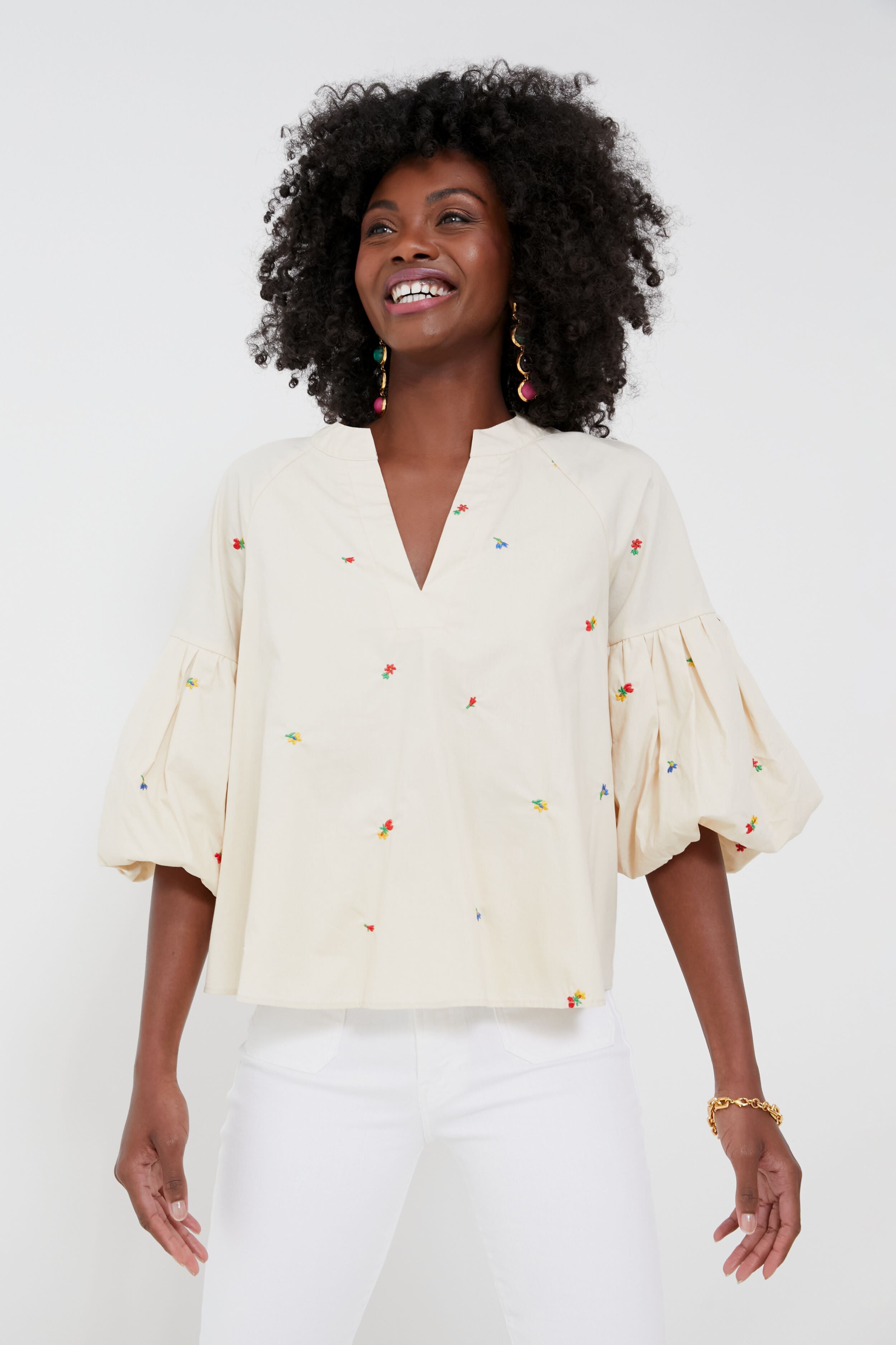 A person wearing a cream-colored, puff-sleeve embroidered blouse with colorful details from Hyacinth House, paired with white pants, stands smiling.