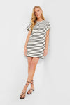 A woman wears the Tuckernuck Black Stripe Lucie Dress, a short-sleeve t-shirt style dress with black and white horizontal stripes, paired with black strappy sandals.