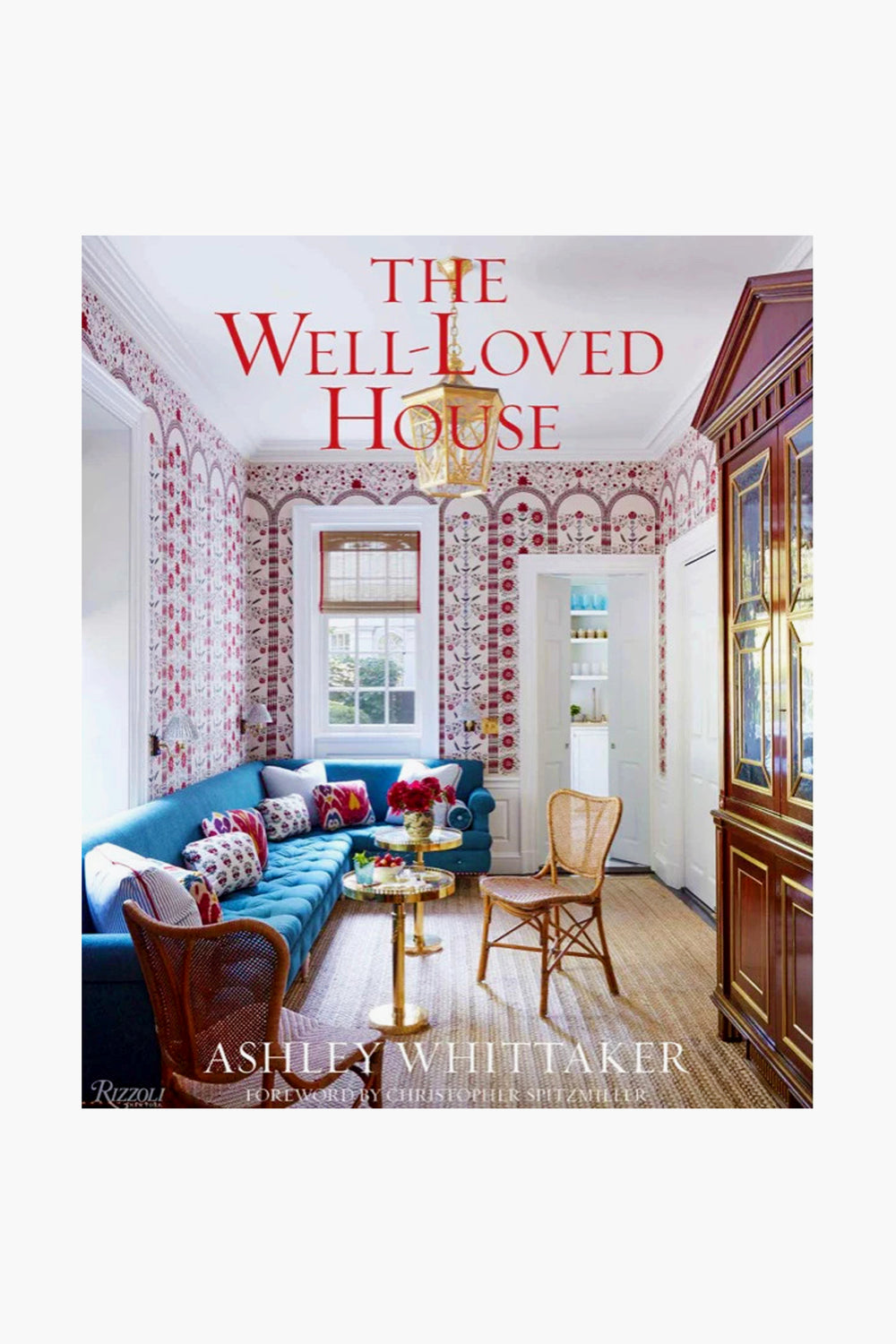Book cover of The Well-Loved House by Ashley Whittaker, published by Rizzoli, featuring a vibrant living room with a blue sofa, patterned accents, wicker chairs, and large windows reflecting classic interior design.