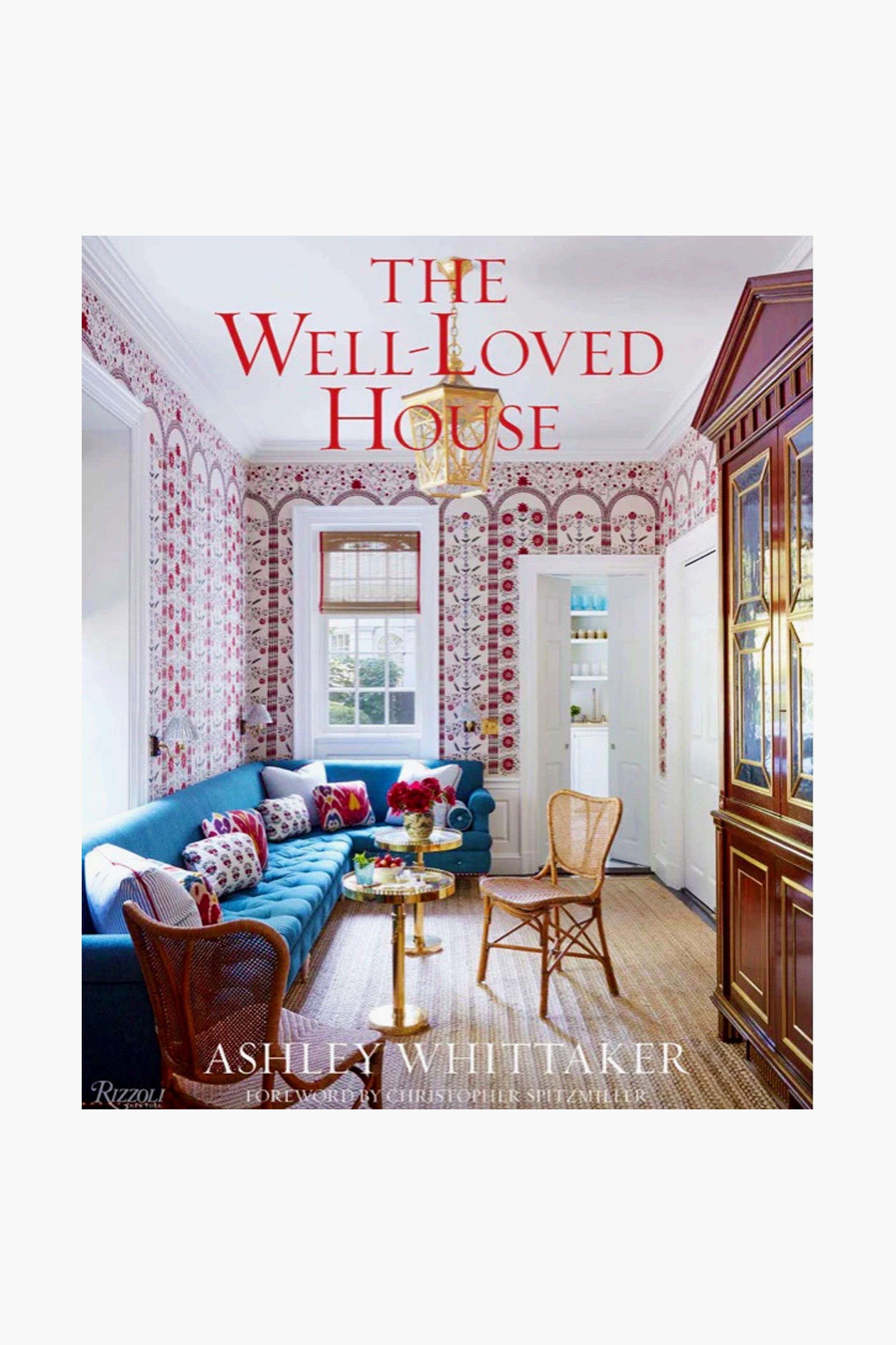 Book cover of The Well-Loved House by Ashley Whittaker, published by Rizzoli, featuring a vibrant living room with a blue sofa, patterned accents, wicker chairs, and large windows reflecting classic interior design.