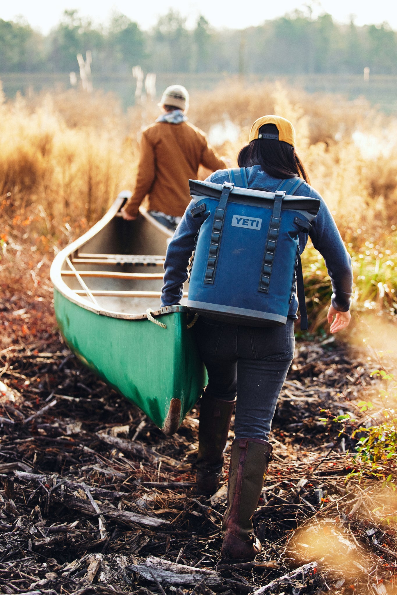 Two people carry a green canoe toward a lake; one wears the classic Navy Hopper M20 Backpack, embodying effortless adventure with high-end, functional design.