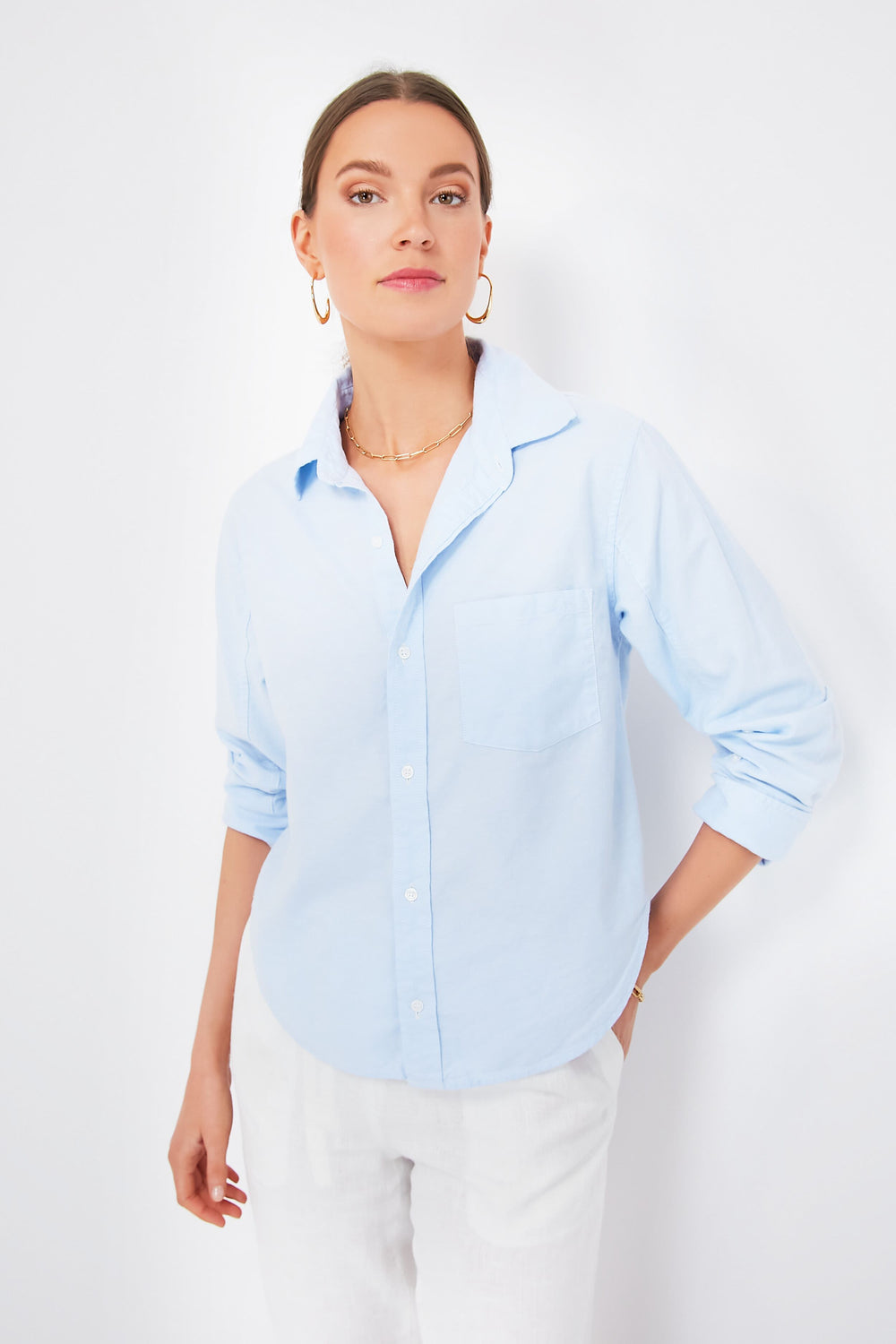 A woman wears a Citizens of Humanity Oxford Blue Kayla Shrunken Shirt with rolled sleeves, paired with white pants, gold hoop earrings, and a necklace—ideal for those who value sustainable fashion.