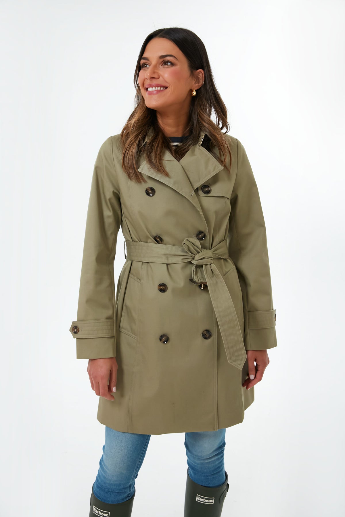 A woman wears a Barbour khaki double-breasted showerproof trench coat with a tie belt, blue jeans, and knee-high green boots.
