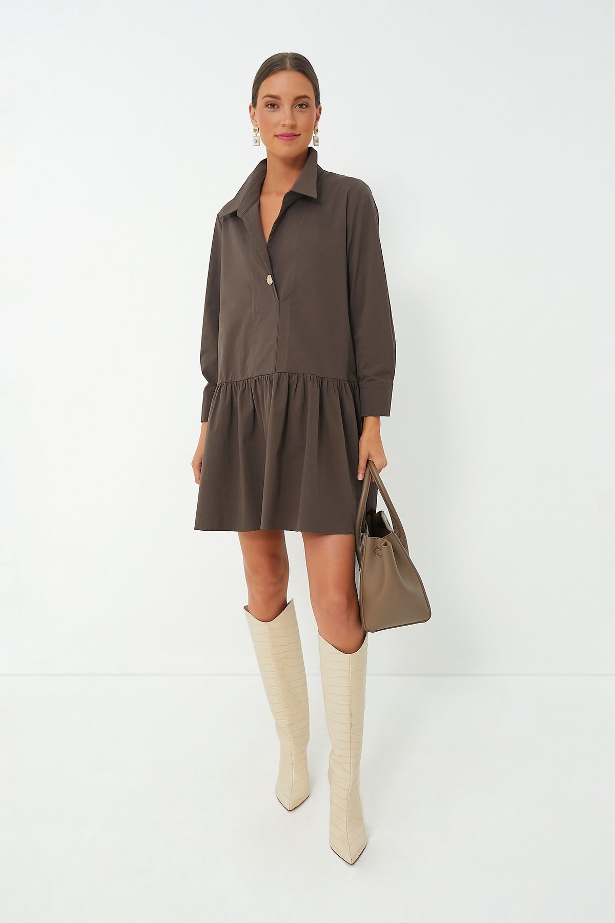 A woman wears a loose brown Pomander Place Cleia Dress with a swing silhouette, cream knee-high boots, and carries a taupe handbag.