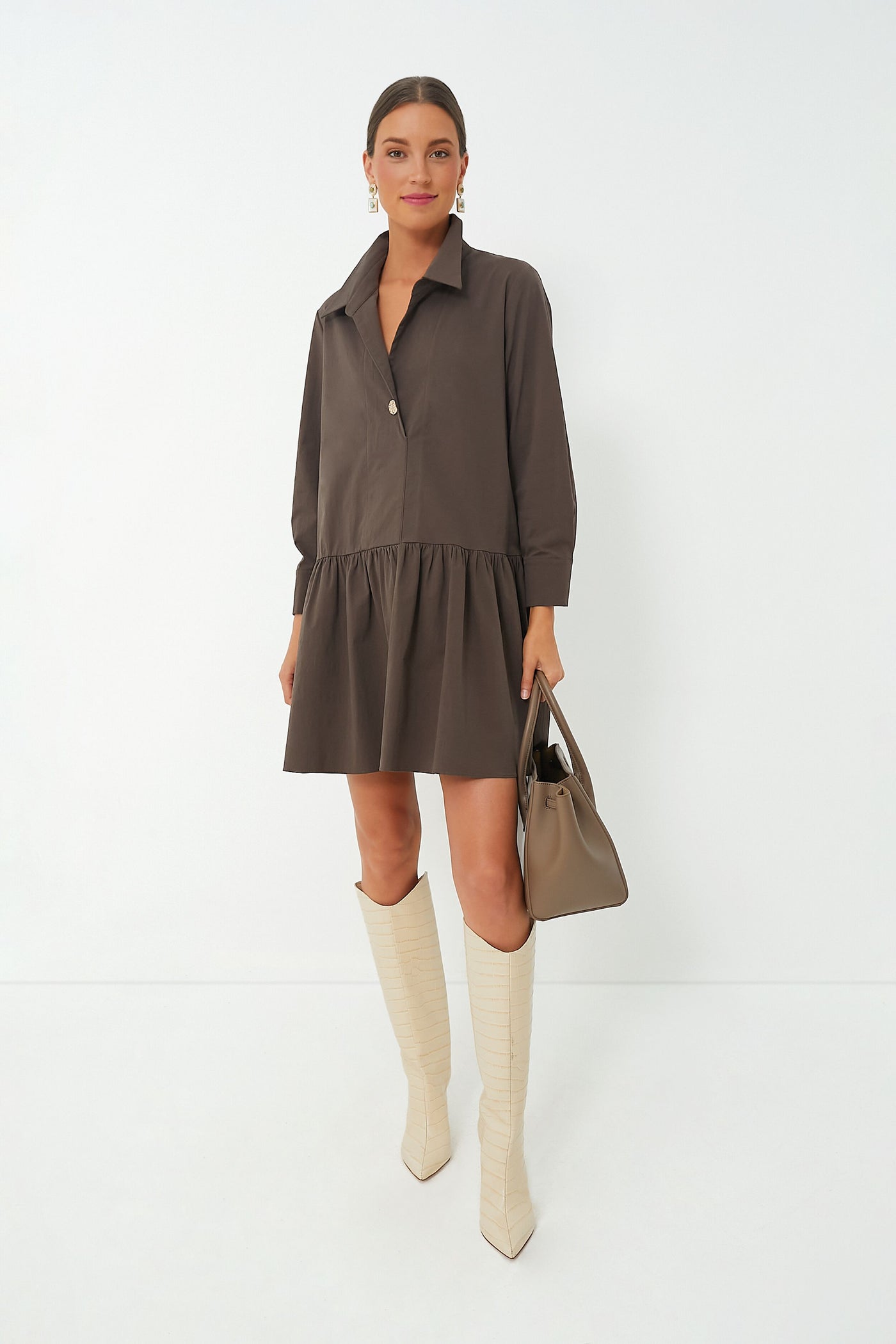 A woman wears a loose brown Pomander Place Cleia Dress with a swing silhouette, cream knee-high boots, and carries a taupe handbag.