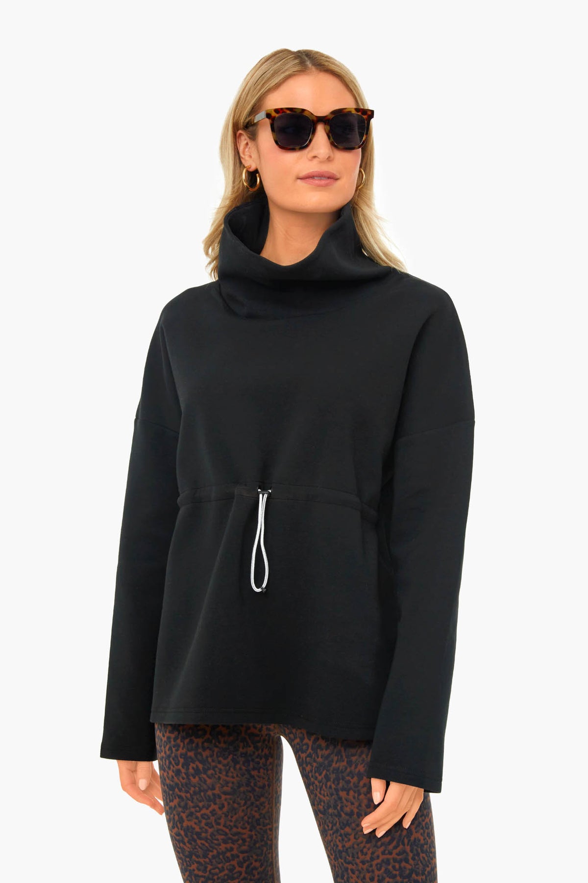 Wearing Varley, a woman in sunglasses showcases a black long-sleeve top with front drawstring, funnel collar, and patterned leggings, embodying performance-fashion with effortless style.