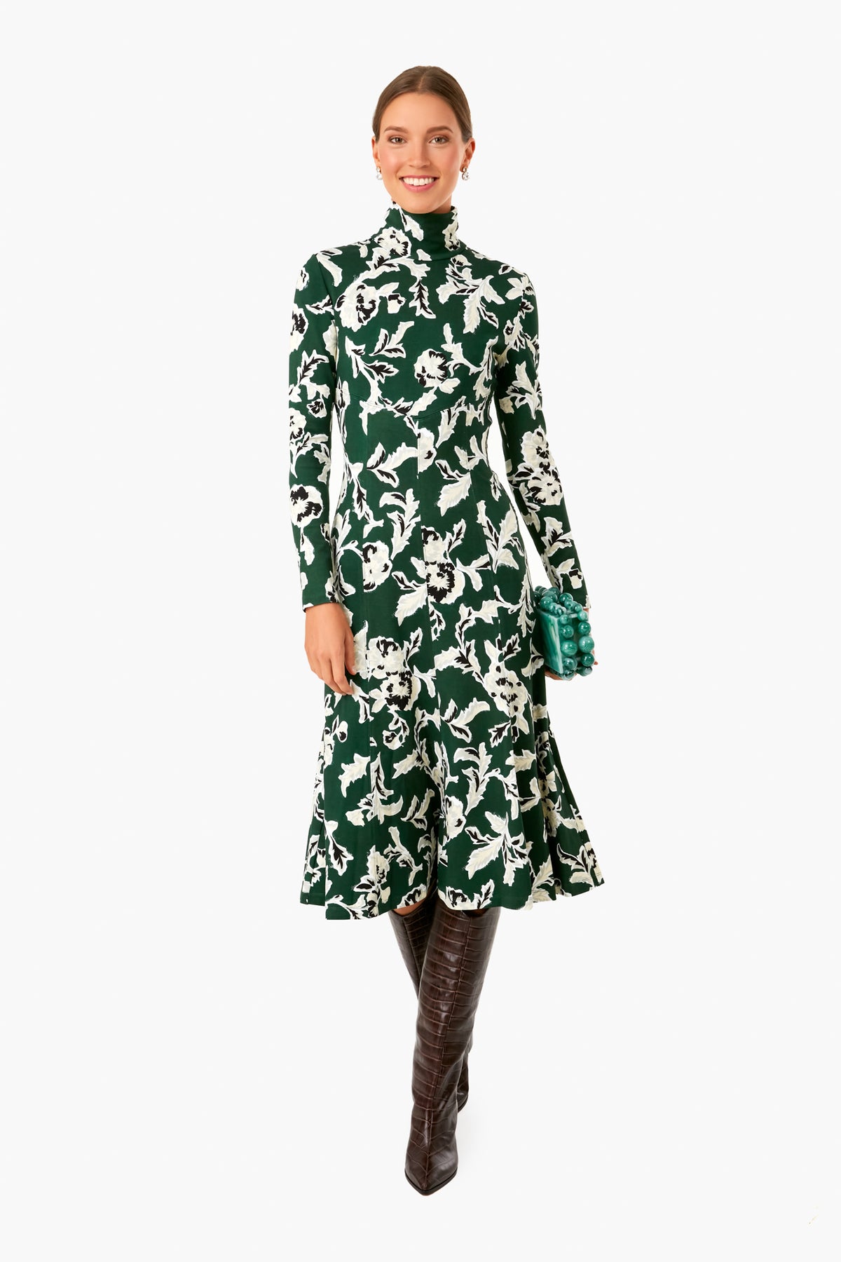 A woman smiles wearing a Tanya Taylor Thea Dress featuring green and white hand-painted prints, long sleeves, and a high neckline, paired with brown knee-high boots and a green clutch.