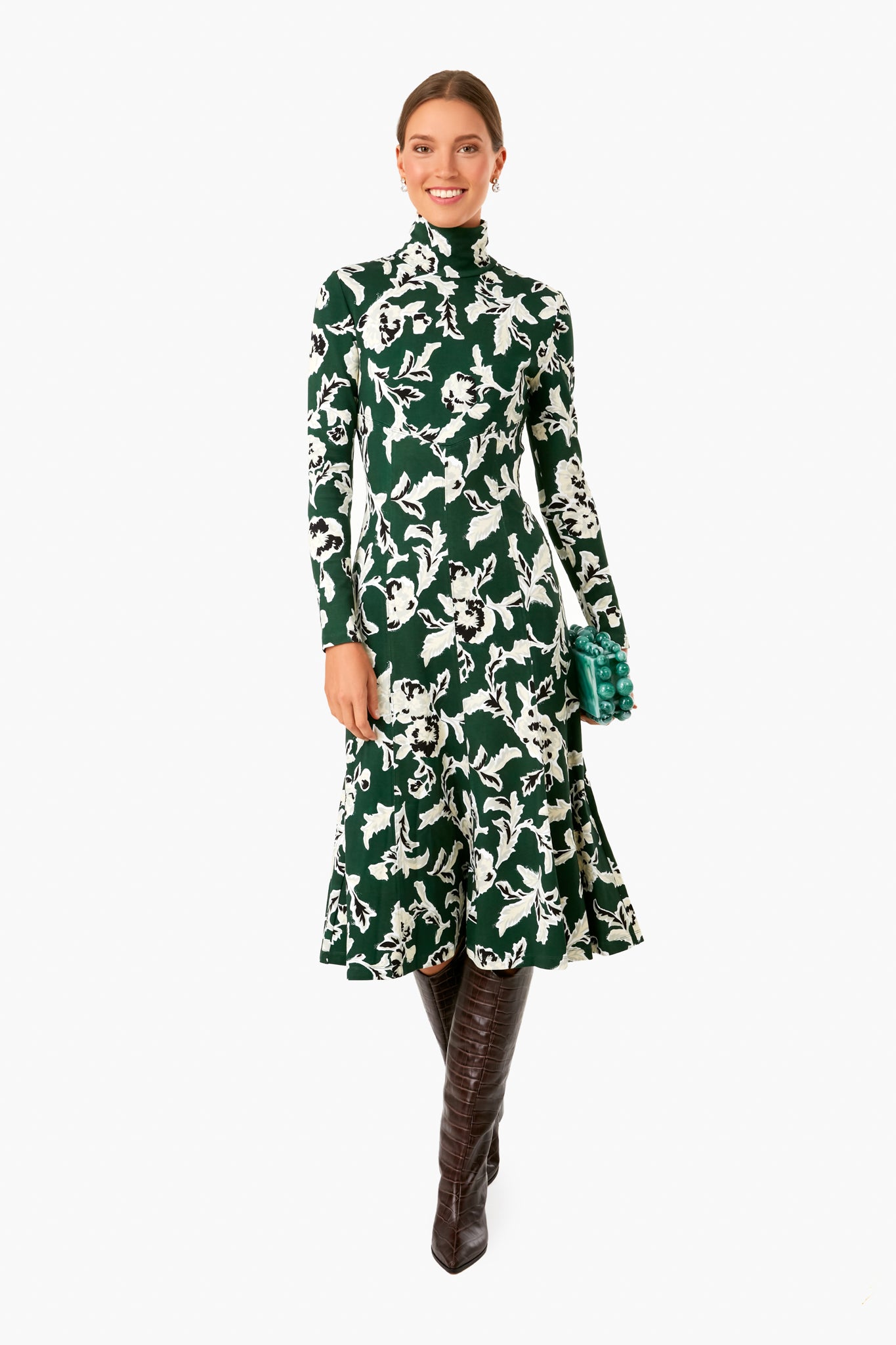 A woman smiles wearing a Tanya Taylor Thea Dress featuring green and white hand-painted prints, long sleeves, and a high neckline, paired with brown knee-high boots and a green clutch.