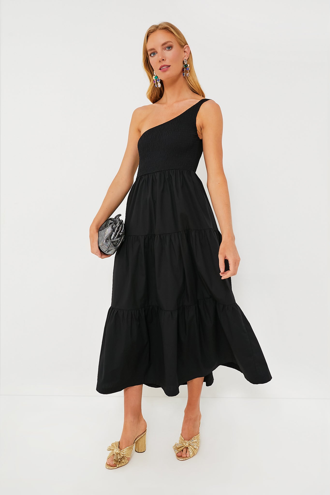A woman in an O.P.T black one-shoulder dress with a tiered midi skirt, styled with gold heeled sandals, statement earrings, and a small black clutch.