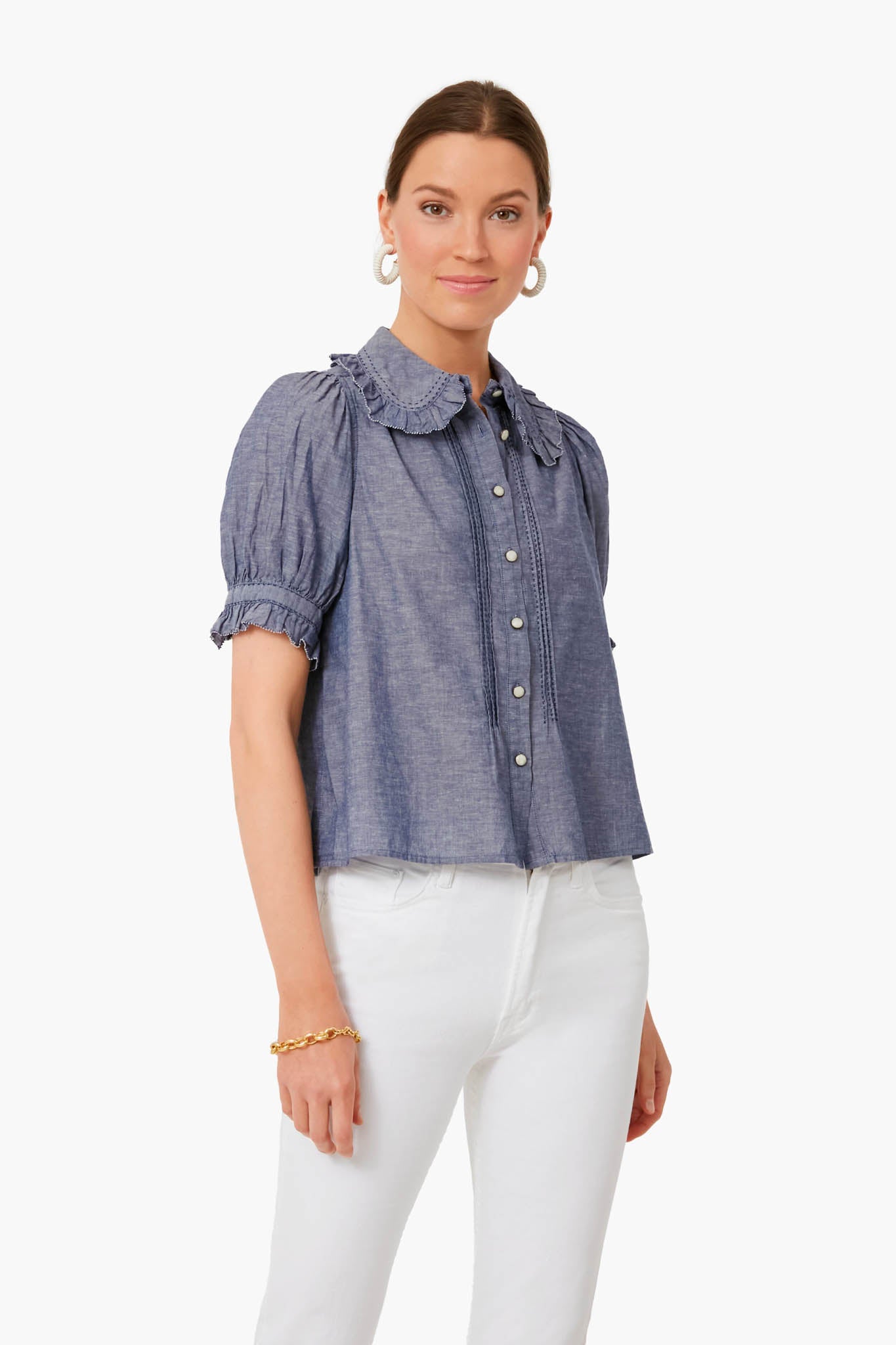 A woman wears THE GREAT.'s navy chambray top with a ruffled collar paired with white pants.