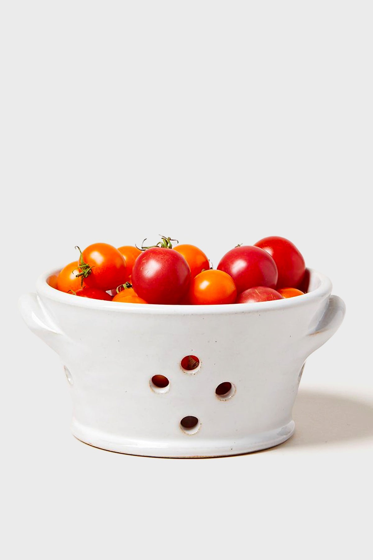 A white ceramic colander from Farmhouse Pottery, Inc brims with red and orange cherry tomatoes, highlighting fresh garden-to-table appeal.