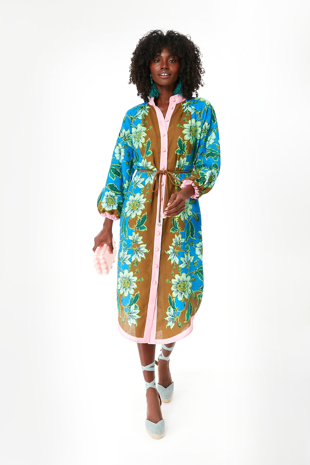 A person wears an Alemais linen midi dress featuring blue, green, and brown floral patterns, pink trim, and a belt, paired with blue lace-up shoes.