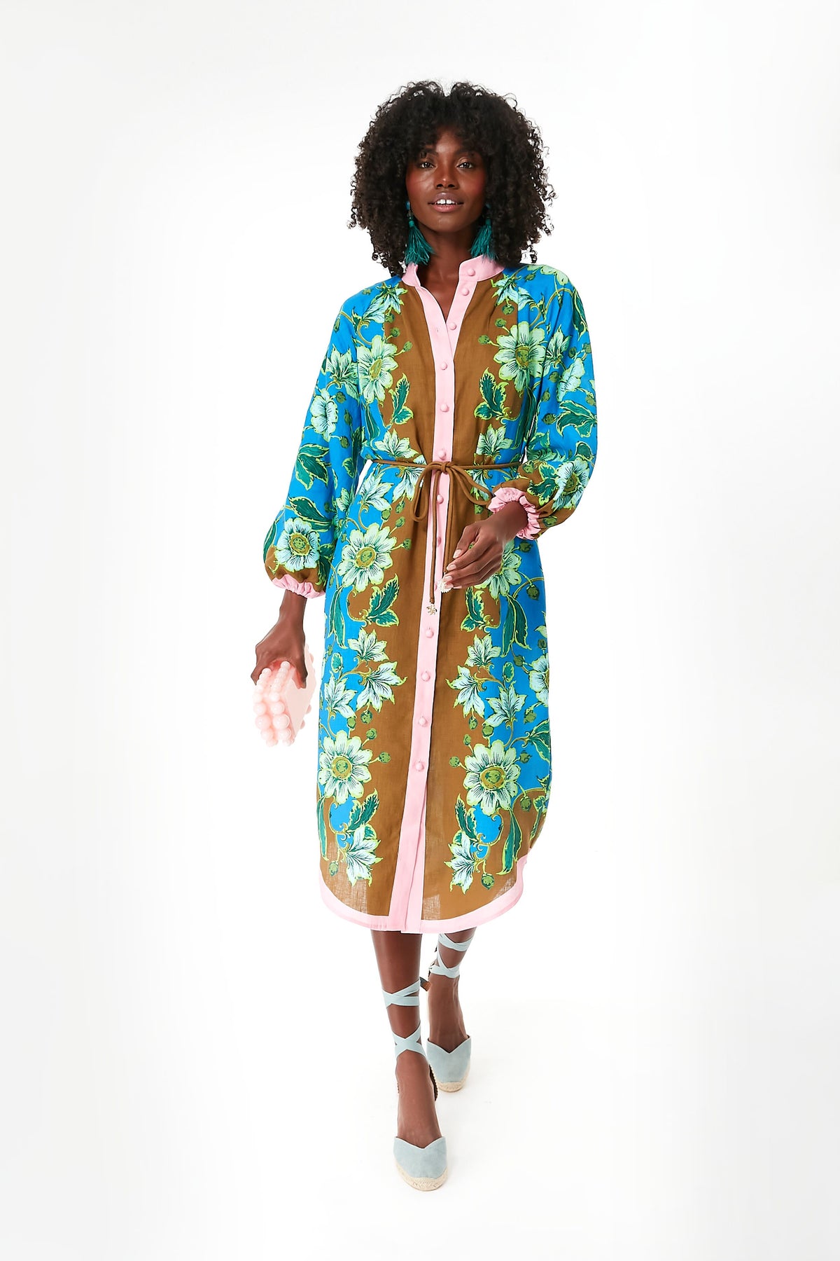 A person wears an Alemais linen midi dress featuring blue, green, and brown floral patterns, pink trim, and a belt, paired with blue lace-up shoes.
