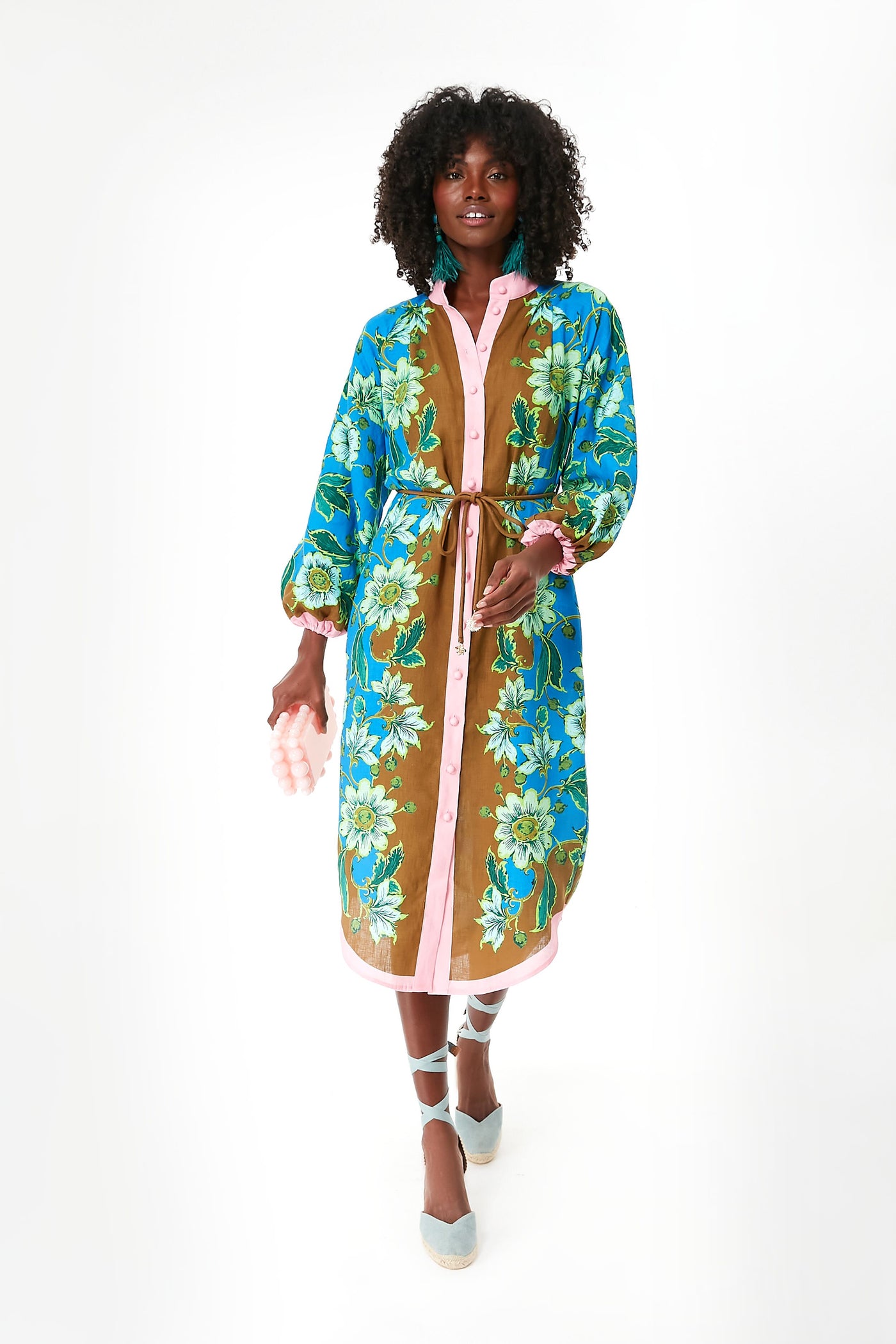 A person wears an Alemais linen midi dress featuring blue, green, and brown floral patterns, pink trim, and a belt, paired with blue lace-up shoes.