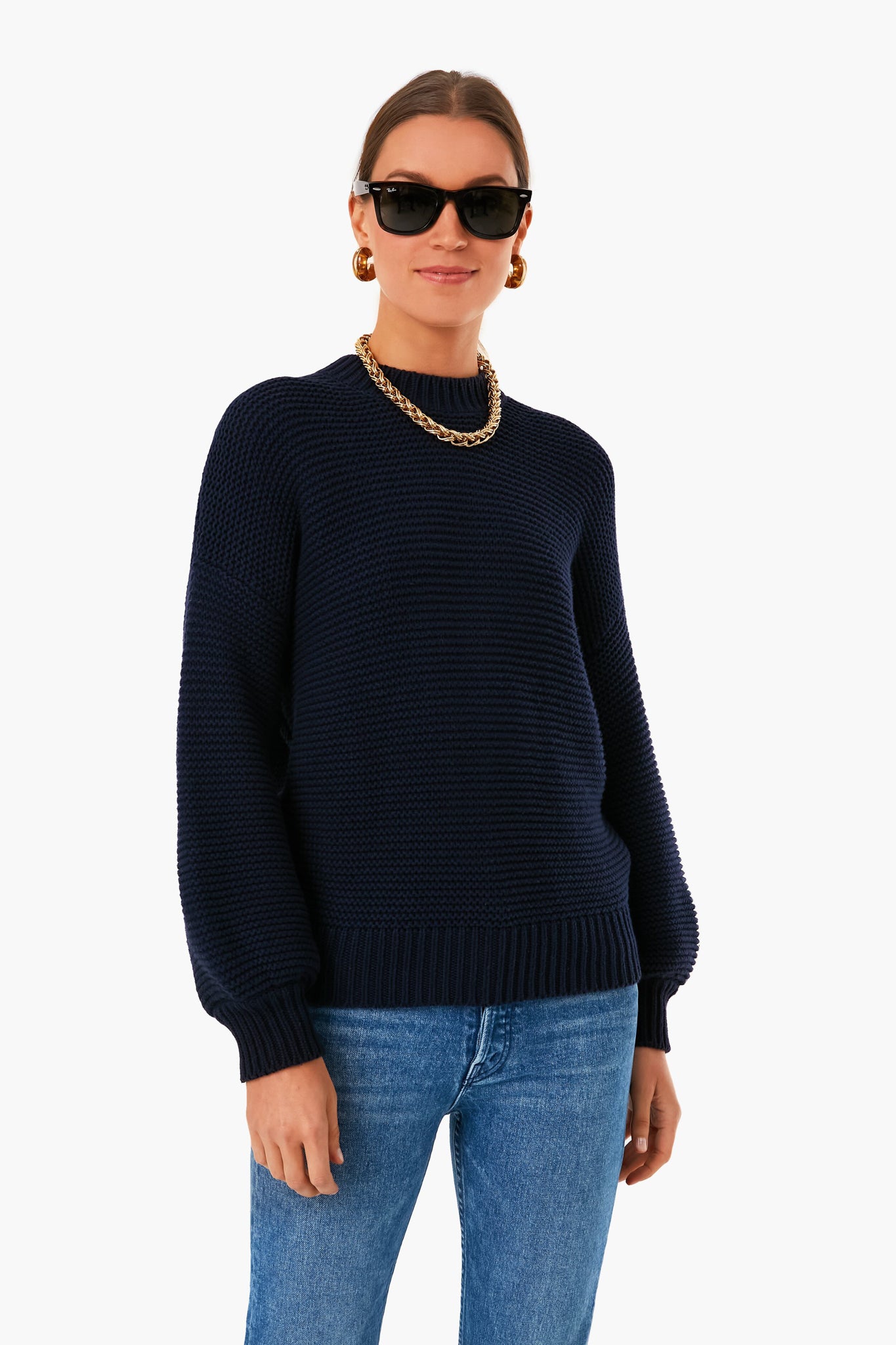 A woman stands confidently in a Dark Navy Button Back Crewneck Sweater by Alex Mill, blue jeans, a gold chain necklace, hoop earrings, and black sunglasses.