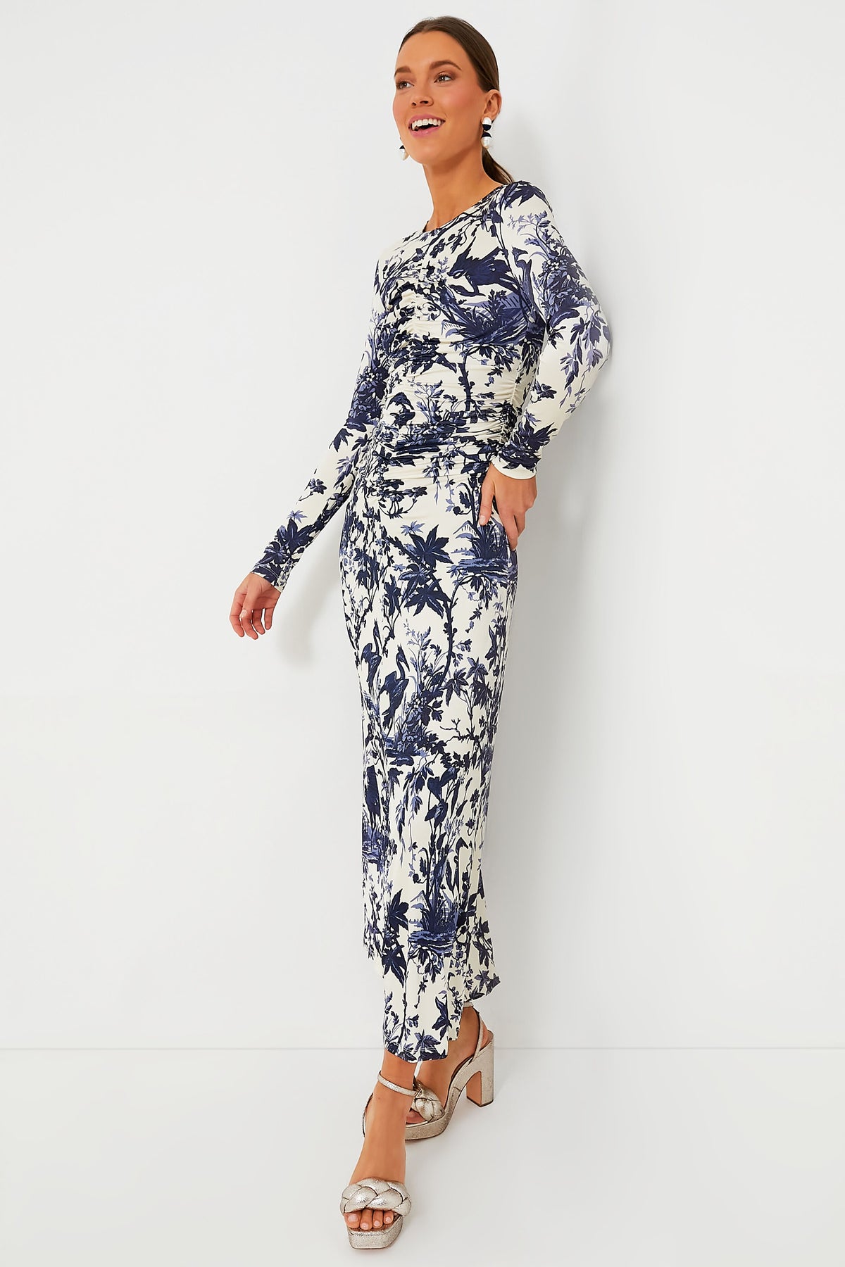 A woman models a Cara Cara long-sleeve, ankle-length white A-line dress with blue botanical print, paired with white heeled sandals and statement earrings.
