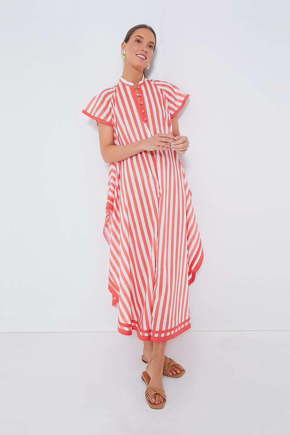 Woman wears the Loretta Caponi Coral Stripes Anouk Dress—a red and white vertically striped, short-sleeve, midi-length cotton caftan with a handkerchief hem—paired with brown flat sandals.