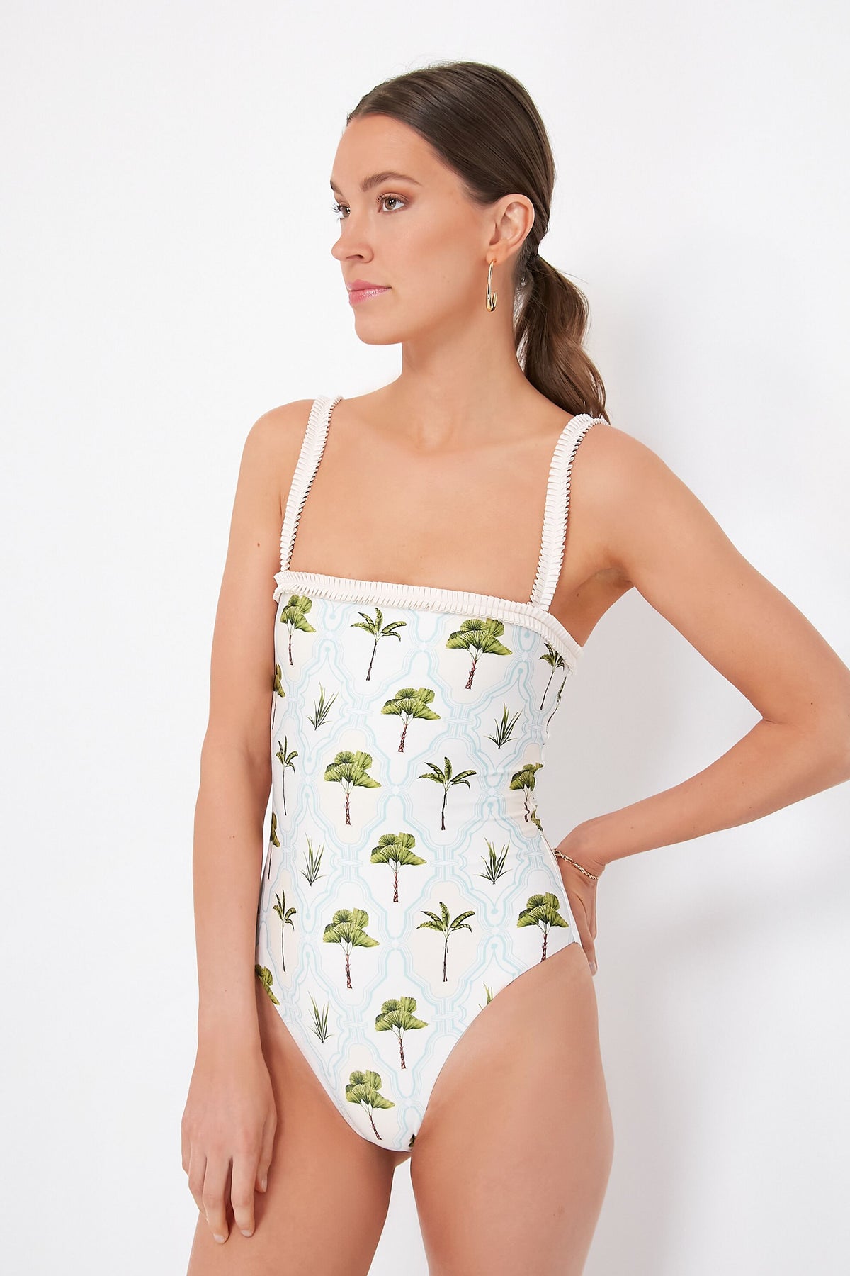 A woman stands confidently with her hand on her hip, wearing an AGUA BY AGUA BENDITA one-piece swimsuit featuring tropical palm tree and plant prints.