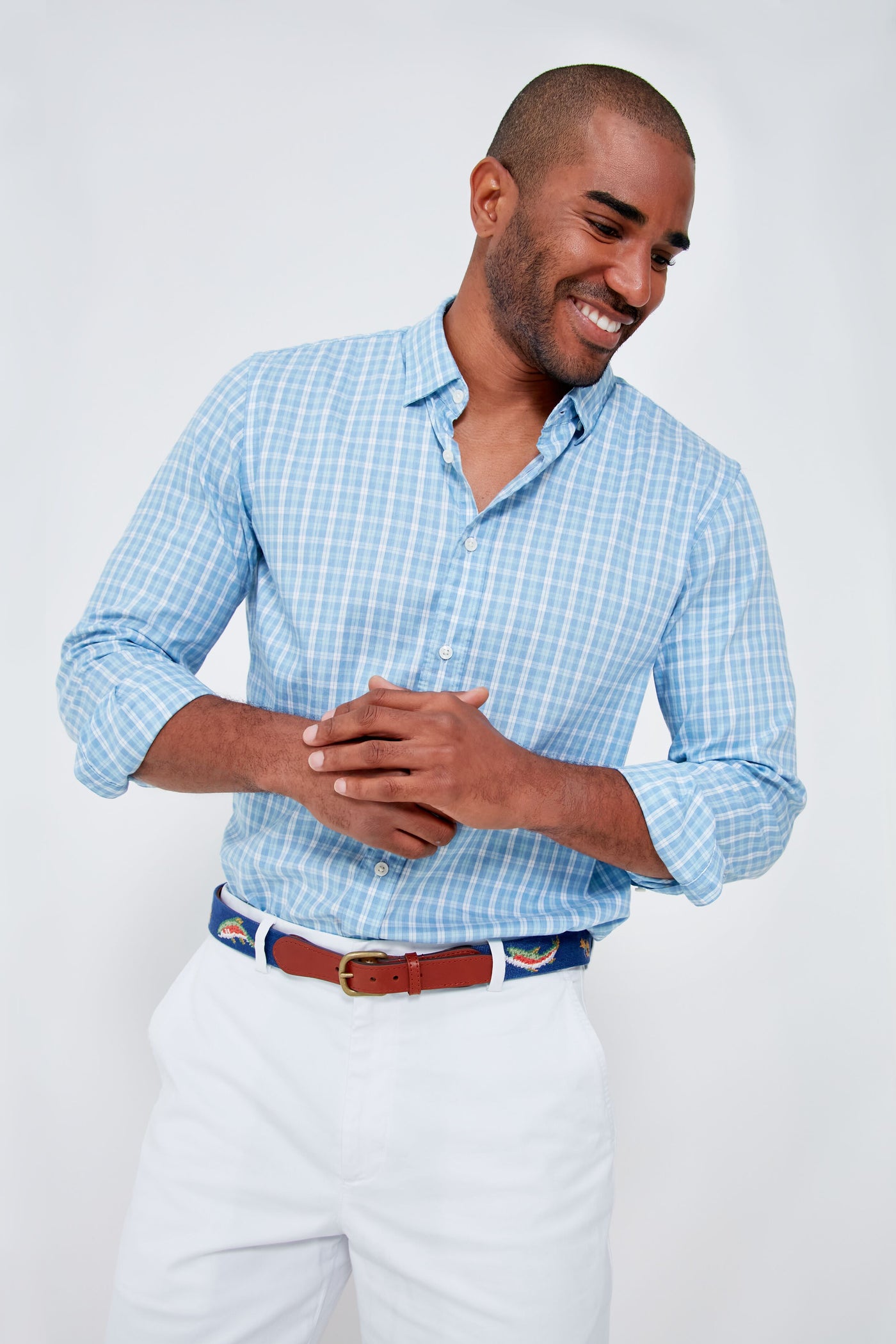 A man wearing a light blue checkered, wrinkle-resistant Supima cotton Faherty shirt and white pants stands smiling with his hands clasped together.