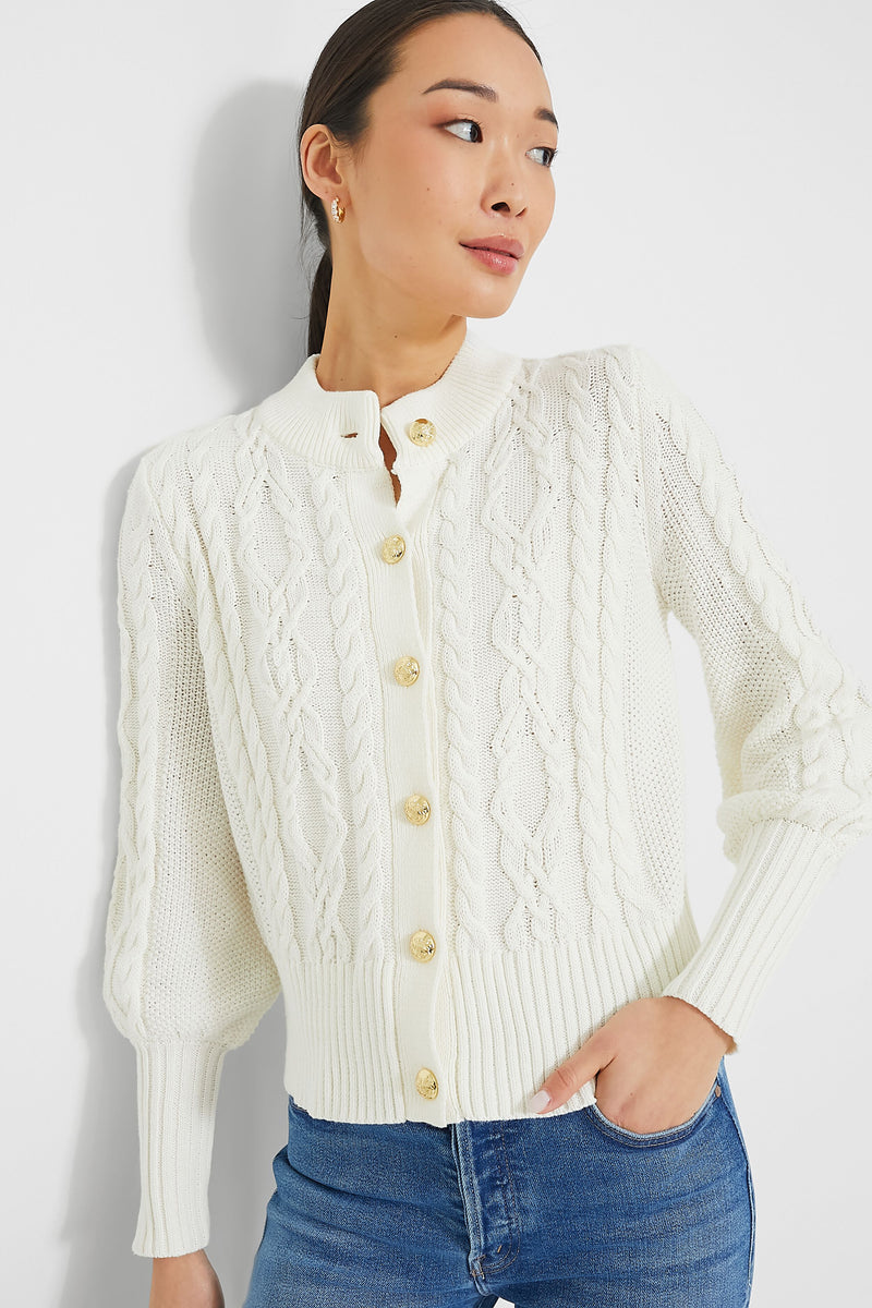 A woman wears a Tuckernuck Ivory Colette Cardigan, a white cable-knit sweater with gold buttons, paired with blue jeans.