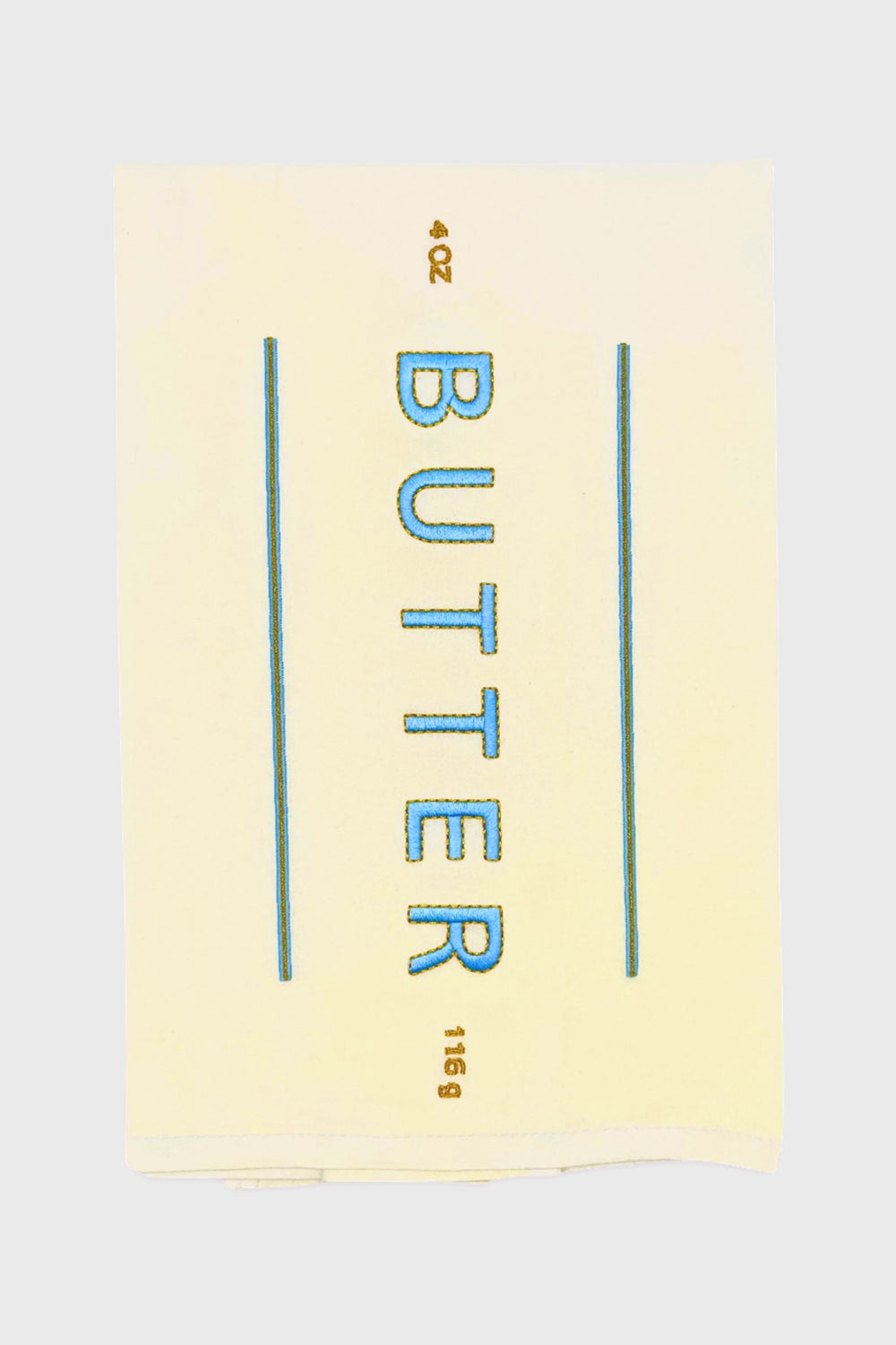 A Lynen Butter Me Up Tea Towel displays a stick of butter labeled "BUTTER" in blue, with 4 OZ and 113g in brown—an embroidered cotton kitchen towel that's perfect for your kitchen.