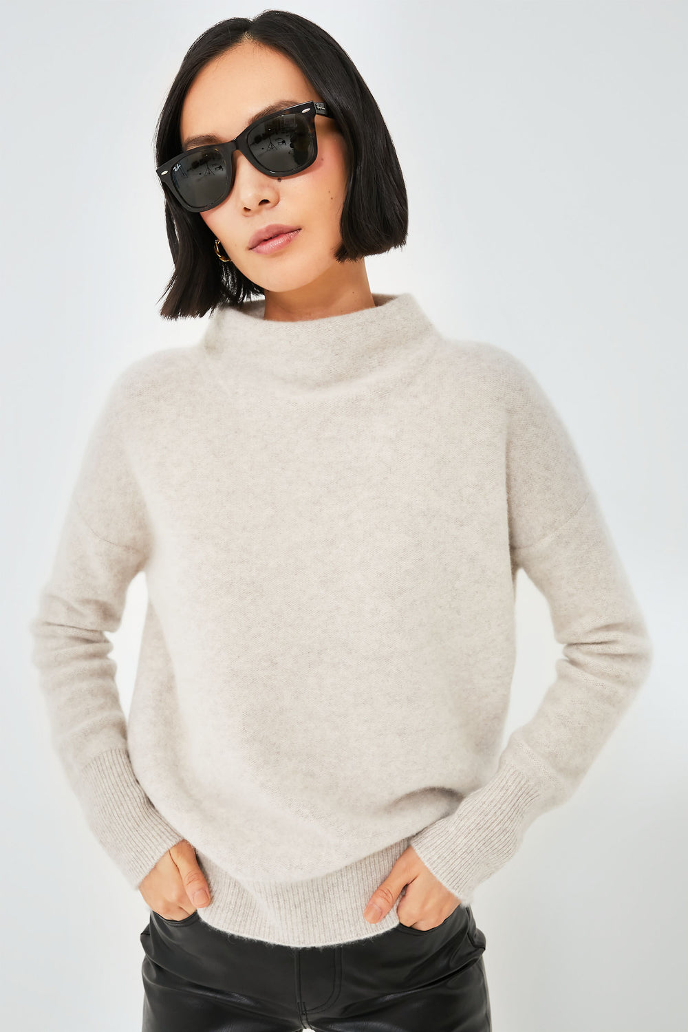 A woman, exuding confidence with hands in pockets, wears black sunglasses and black leather pants paired with a Vince light beige cashmere sweater in a relaxed fit.