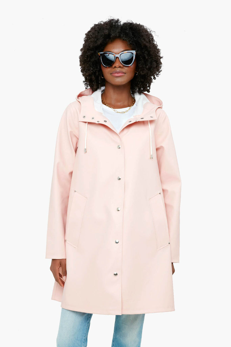 A woman wearing a light pink Stutterheim handmade rubberized cotton raincoat, sunglasses, a white top, and blue jeans stands confidently.