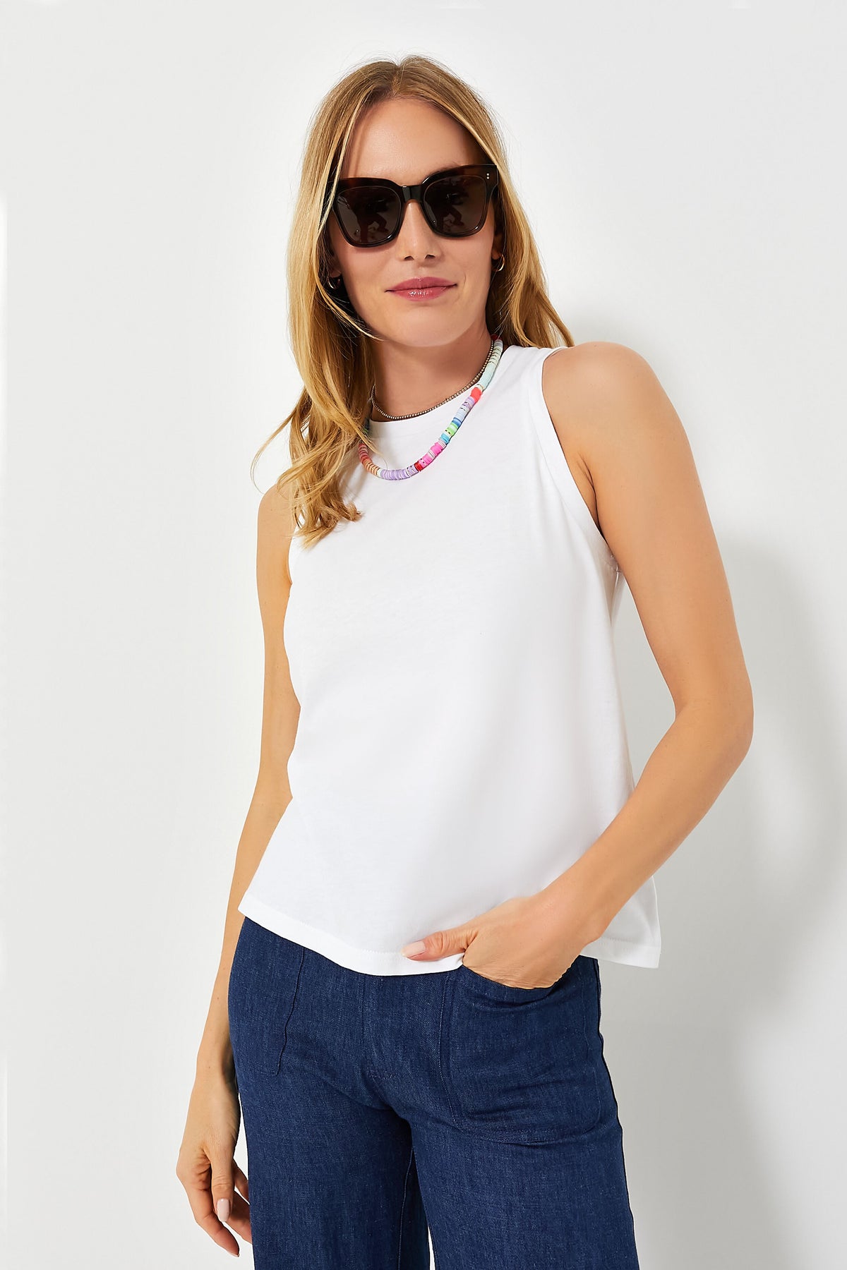 Woman wears a sleeveless white Pomander Place Anthos Shell, dark sunglasses, blue pants, and a colorful beaded necklace.