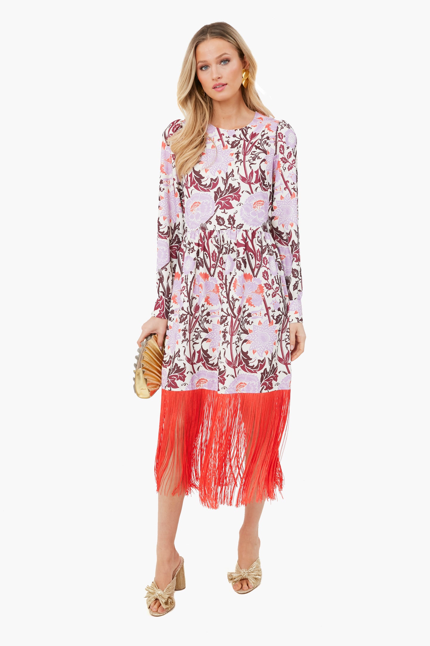 A woman in the Tuckernuck Lonestar Lavender Aerin Dress, a long-sleeve floral style with red fringe at the hem, carries a gold clutch and wears gold heeled sandals for a rustic-chic flair.
