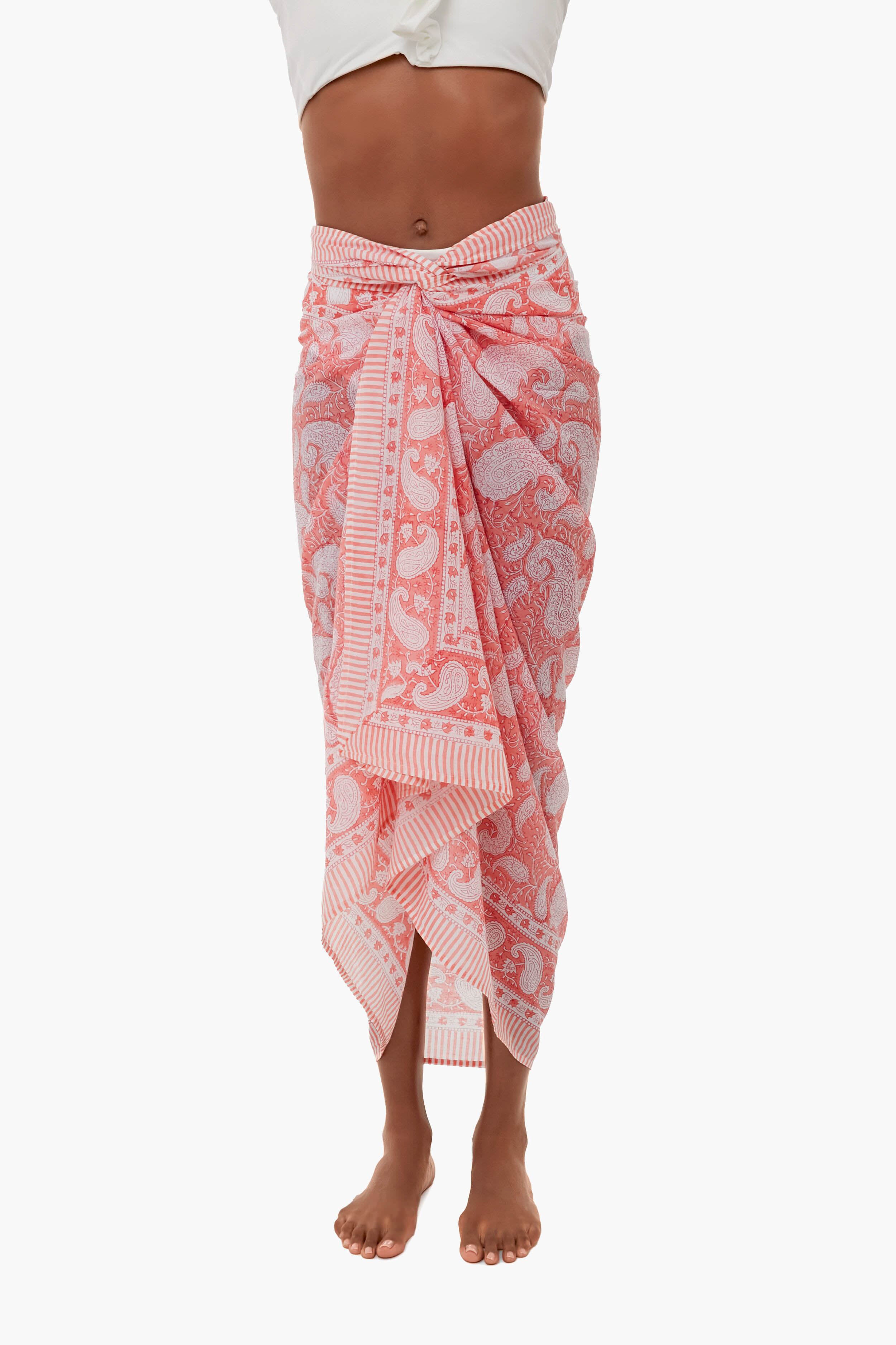 A person stands barefoot in a Sunshine Tienda x Ashley Brooke Pink Sunset Sarong, featuring pink and white patterns, tied at the waist—a perfect lightweight cotton beachwear essential.