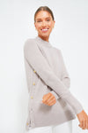 A woman smiles warmly in a light gray Tuckernuck Bexley sweater with gold side buttons, highlighting the stylish wool crewneck.