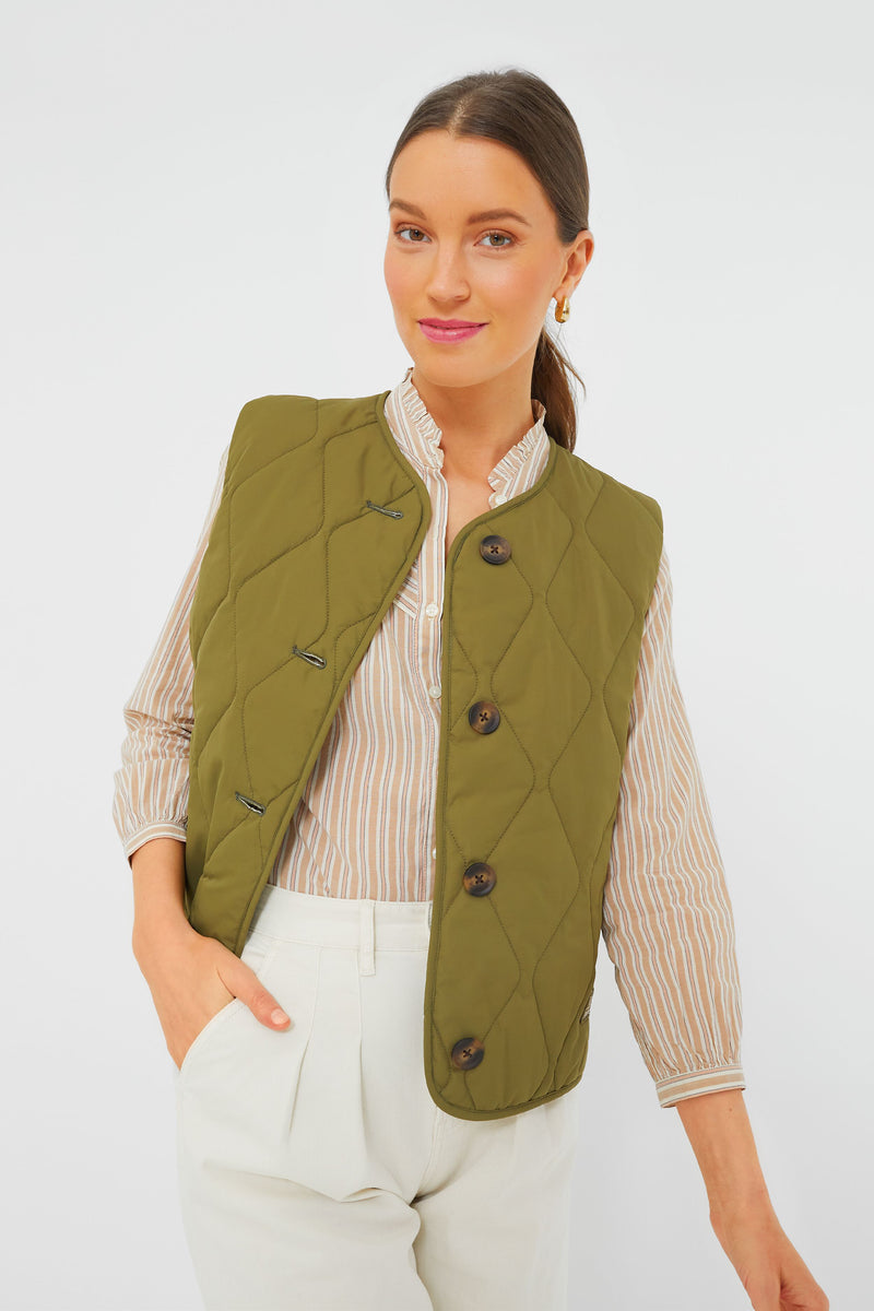 A woman wears a Barbour olive green gilet over a striped shirt with beige trousers, posing with one hand in her pocket. This Barbour outerwear adds a classic touch to any outfit.