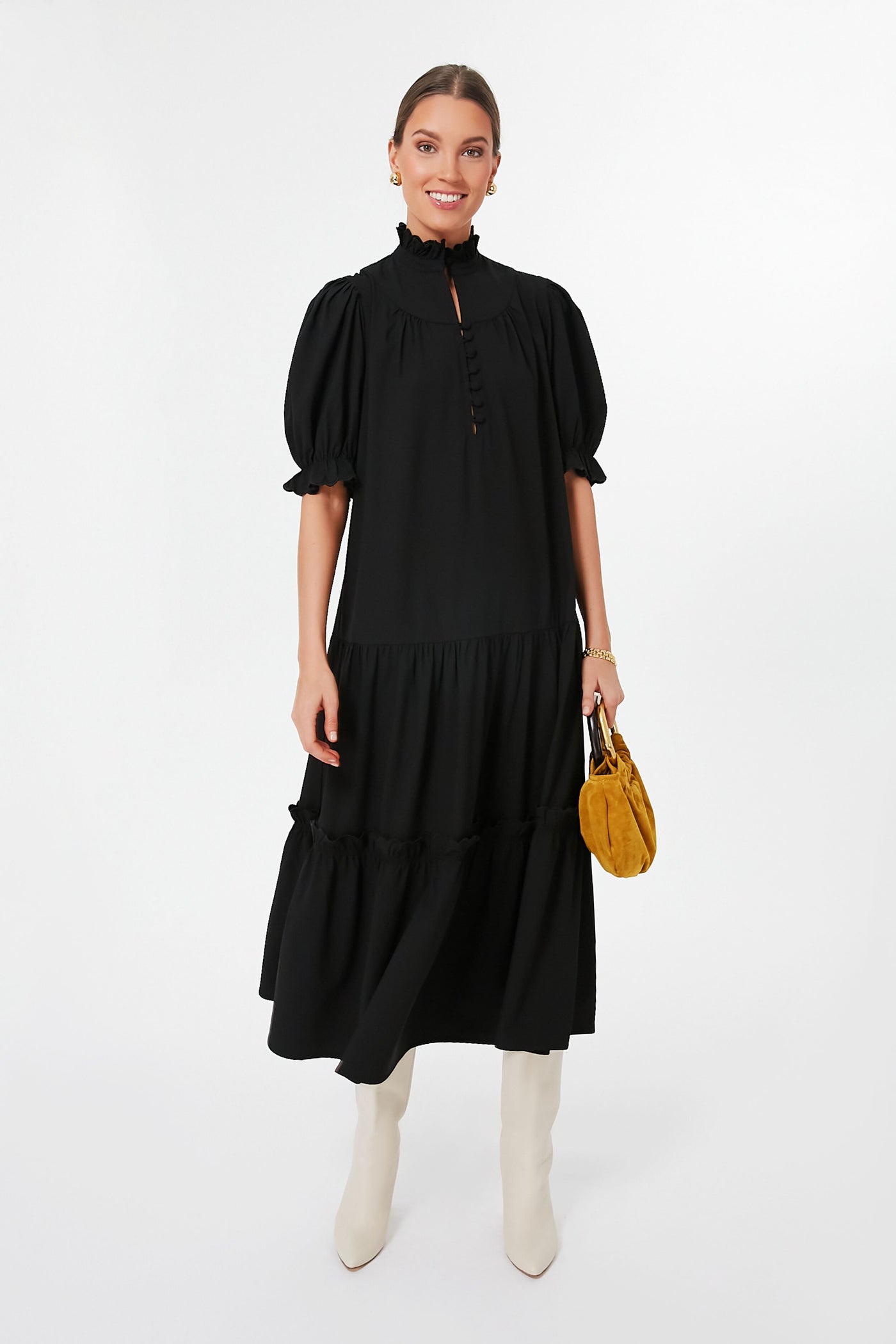 A woman smiles in a long black puff sleeve dress with a ruffle stand collar from Pomander Place, styled with white knee-high boots and a small mustard yellow handbag.