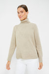 A woman stands poised in a simple setting, dressed in a beige Barbour turtleneck sweater with a diamond trellis knit and white pants.