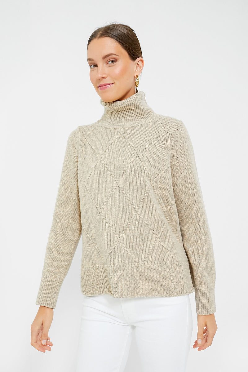 A woman stands poised in a simple setting, dressed in a beige Barbour turtleneck sweater with a diamond trellis knit and white pants.