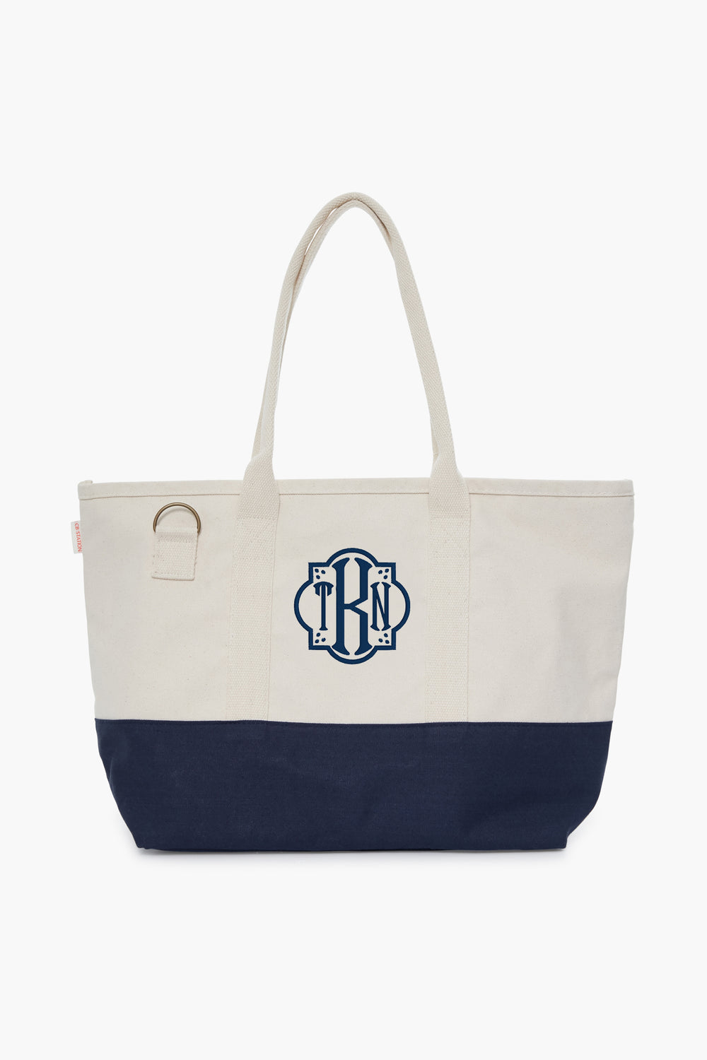 The CB Station two-tone cotton canvas bag features a cream top, navy bottom, two handles, side ring, and blue monogram on the front—an eco-friendly choice for daily use.