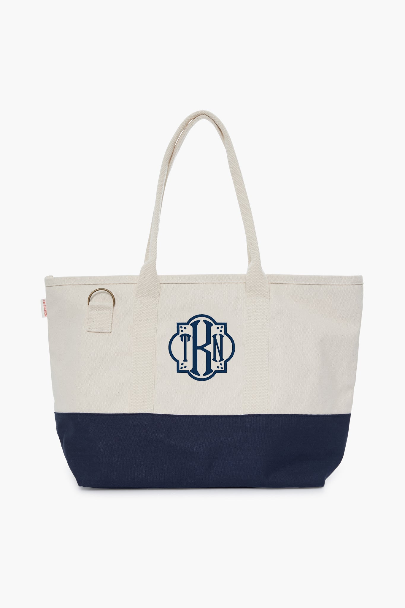 The CB Station two-tone cotton canvas bag features a cream top, navy bottom, two handles, side ring, and blue monogram on the front—an eco-friendly choice for daily use.