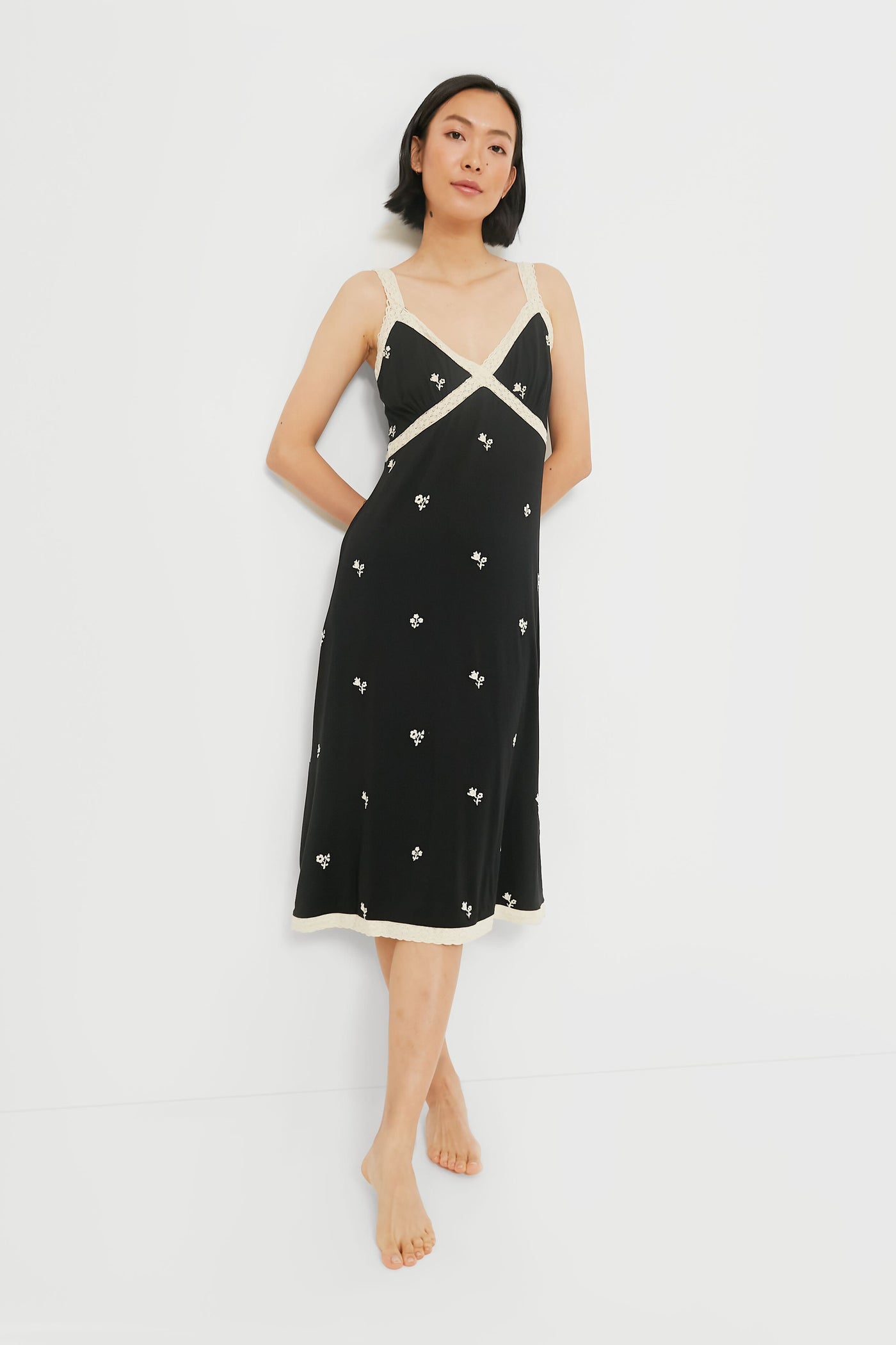 A woman stands barefoot wearing a Sea New York black floral slip dress with white trim and small white floral patterns.