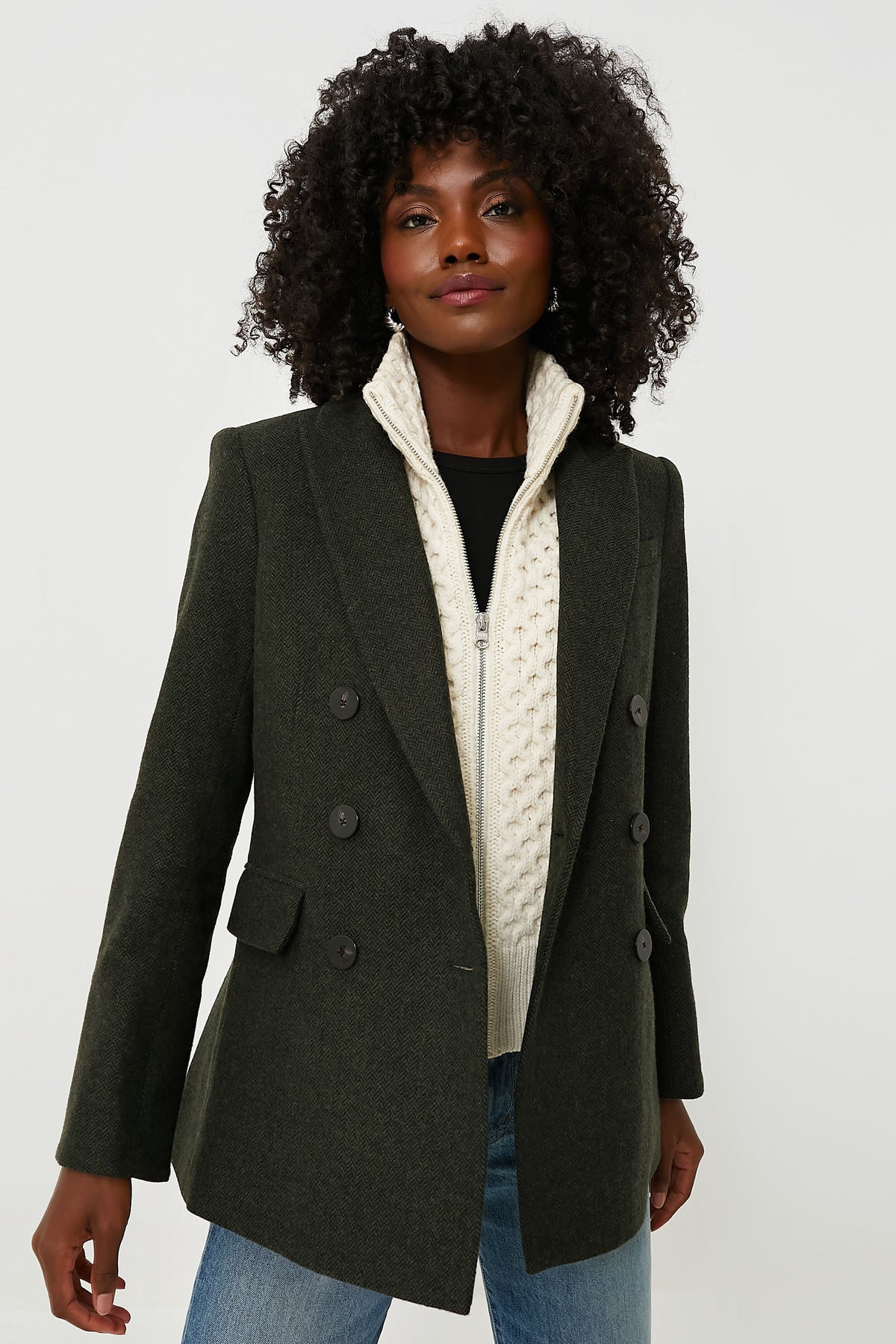 A woman wears a Veronica Beard dark green wool double-breasted blazer over a cream cable knit dickey and blue jeans.
