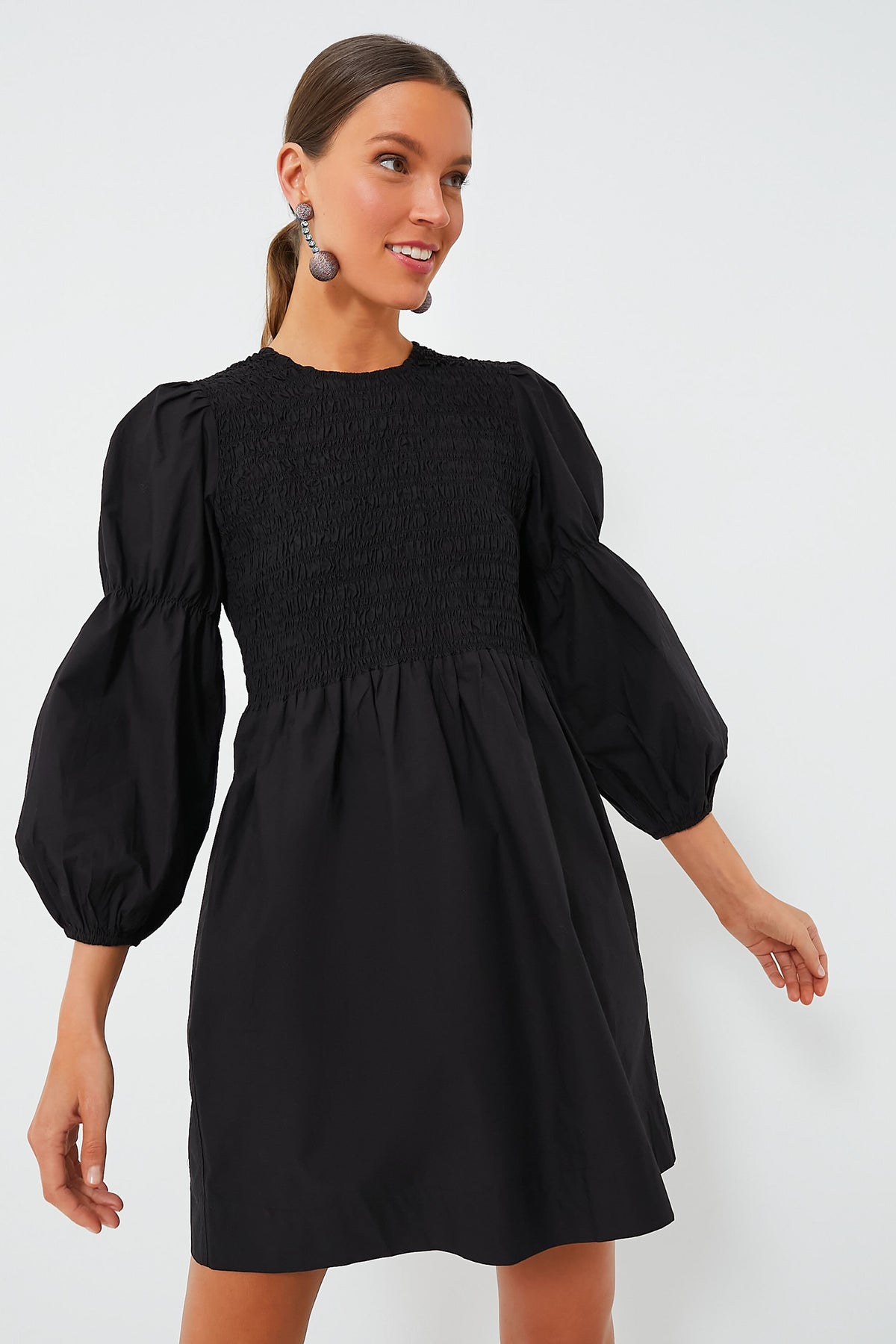A woman wears a GANNI Black Cotton Poplin Smock Dress with puffed three-quarter sleeves. This mini dress is made from organic cotton, combining style and comfort.