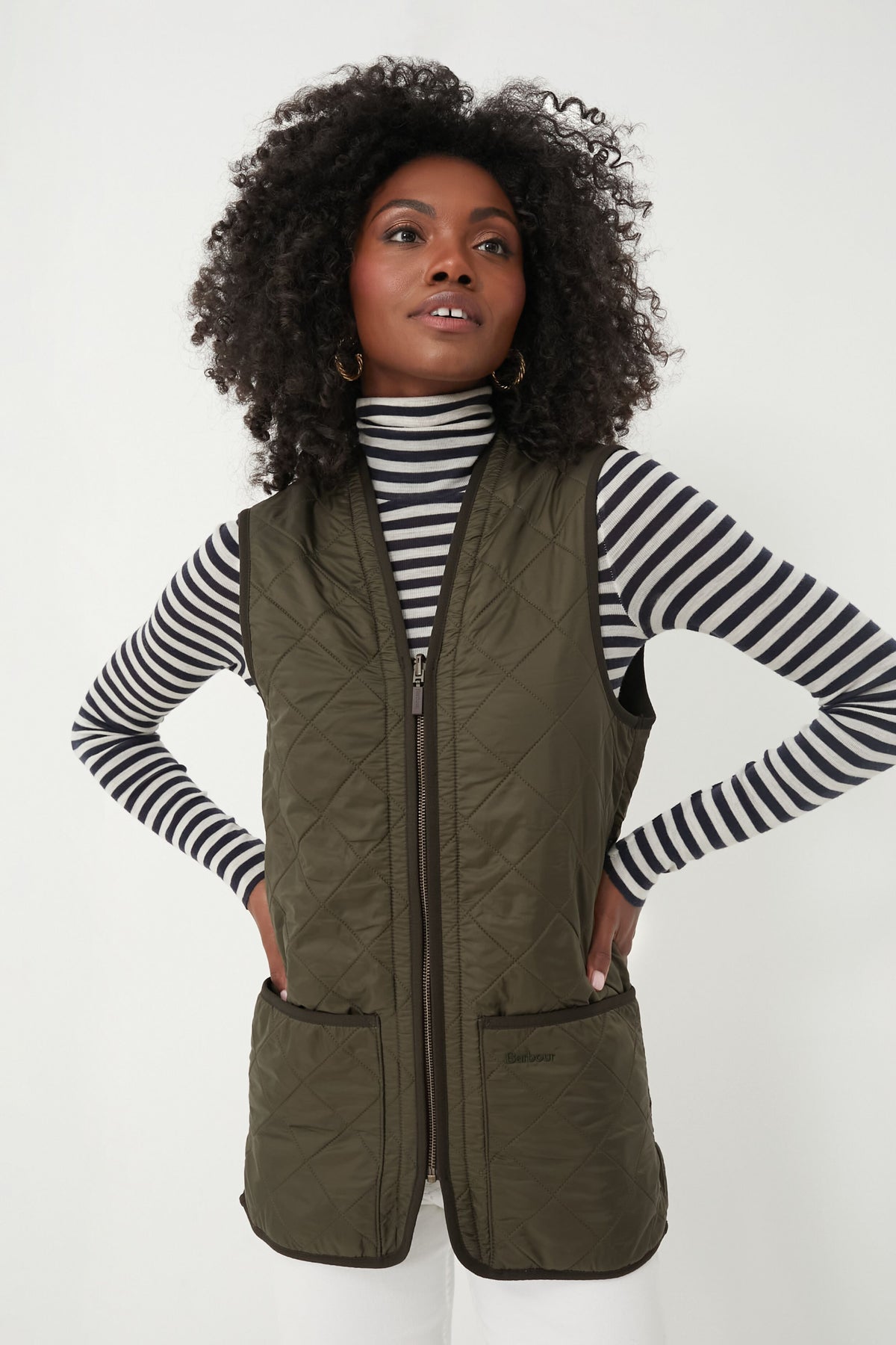 A woman stands with her hands on her hips, wearing a black-and-white striped turtleneck and a Barbour olive Polarquilt Waistcoat.