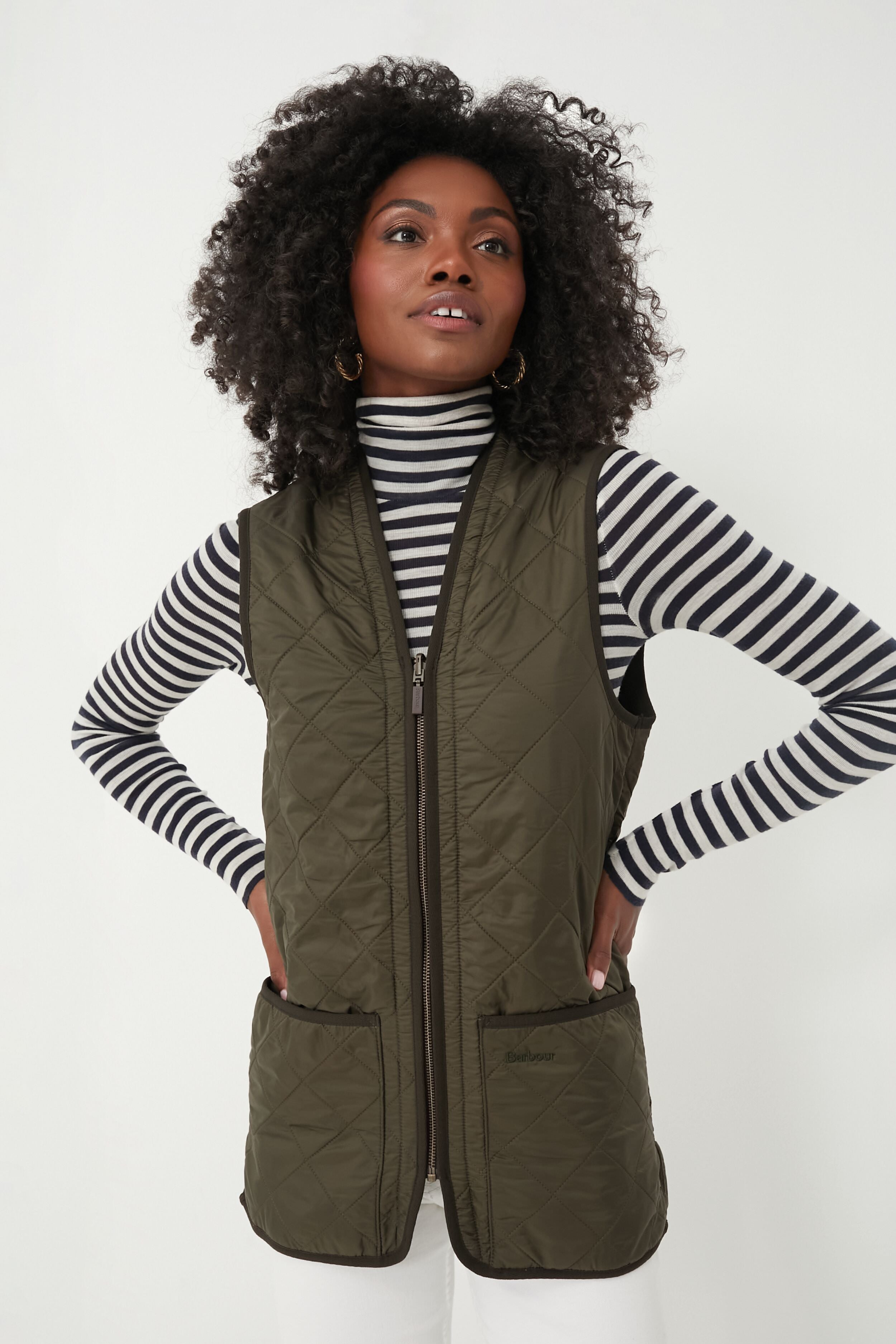 A woman stands with her hands on her hips, wearing a black-and-white striped turtleneck and a Barbour olive Polarquilt Waistcoat.