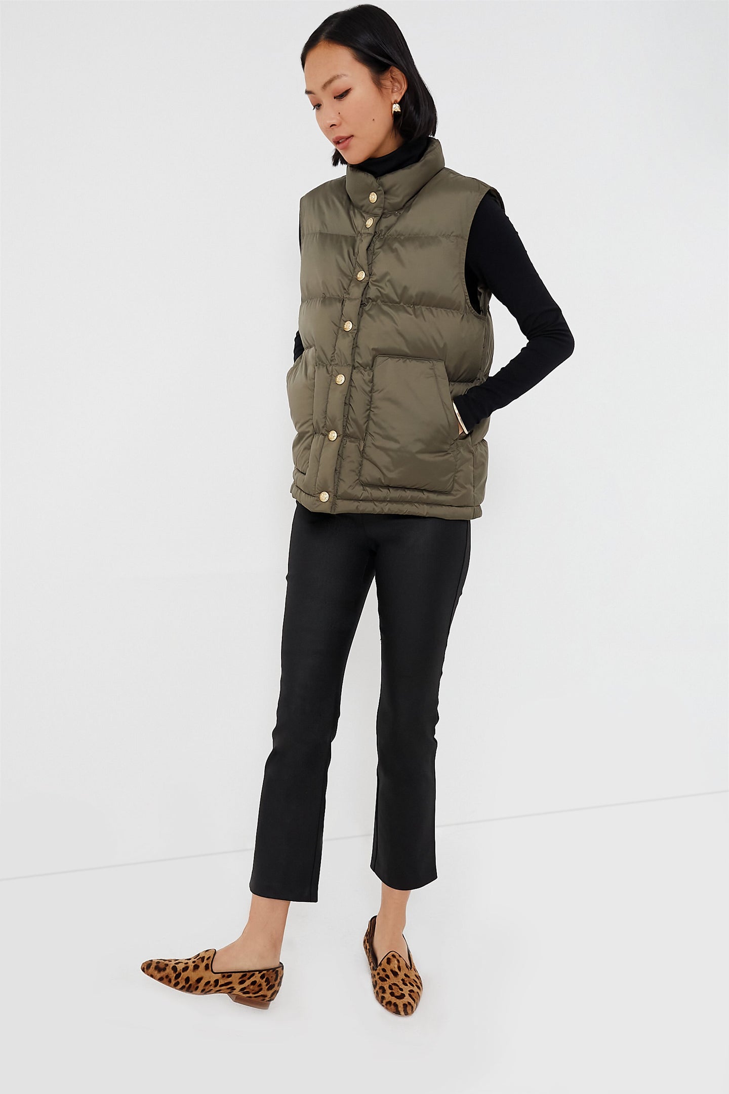 A woman poses against a simple backdrop in Tuckernuck outerwear: an olive green puffer vest with oversized pockets, paired with a black long-sleeve top, black flared pants, and leopard print slip-on shoes.