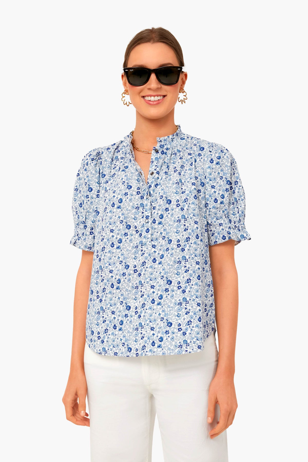 A woman wears Apiece Apart black sunglasses, gold earrings, and a light blue floral puff shoulder top with ruffled collar, paired with white pants—all crafted from soft organic cotton for effortless style.