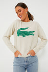 Woman wearing a cream cashmere sweater with a large green alligator graphic on the front from the Bandier x Lacoste collection, paired with light blue jeans.