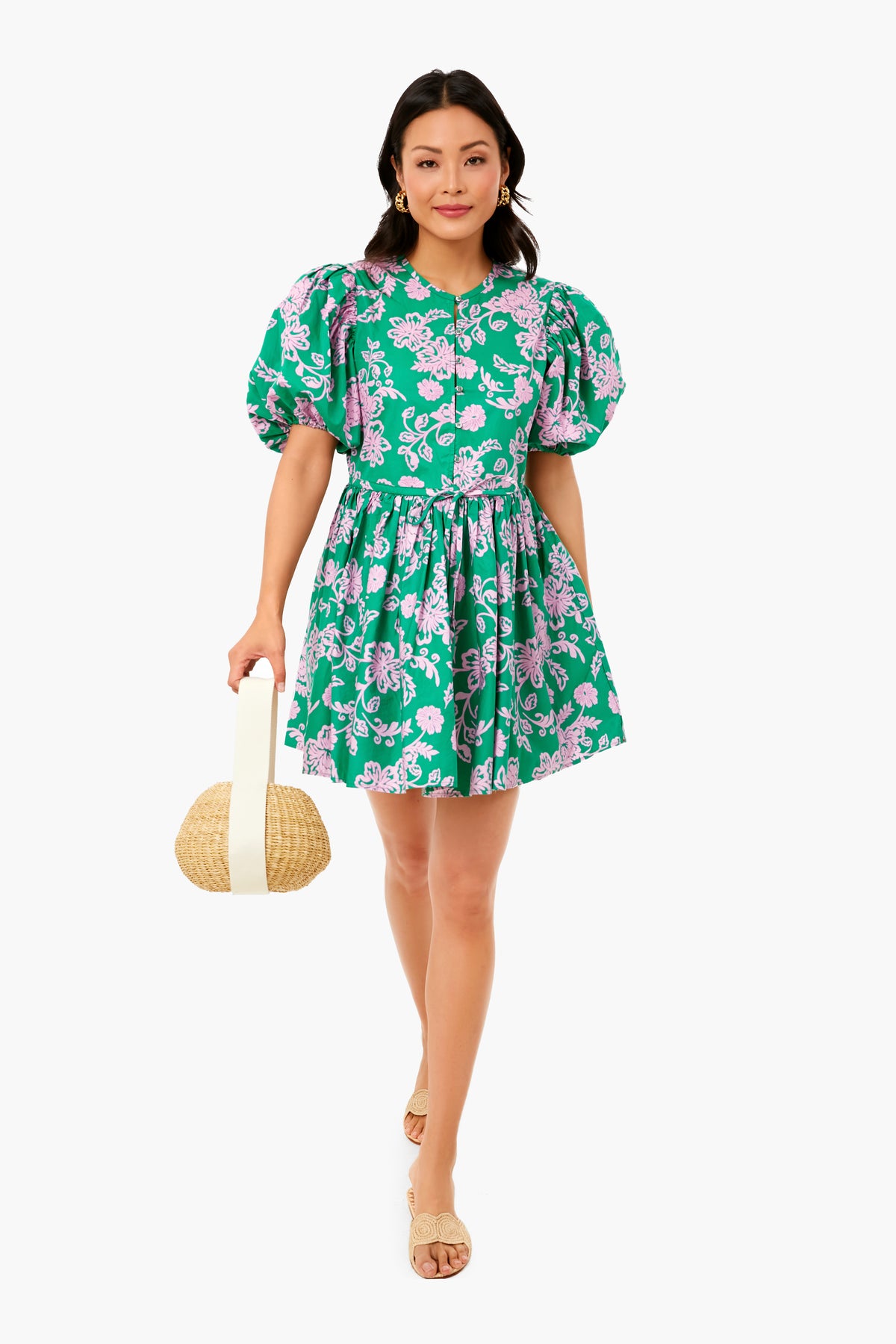 A woman in a XiRENA Caprisyn Green Aurie mini dress with pink floral pattern and puff sleeves, paired with beige sandals, holds a round straw handbag.
