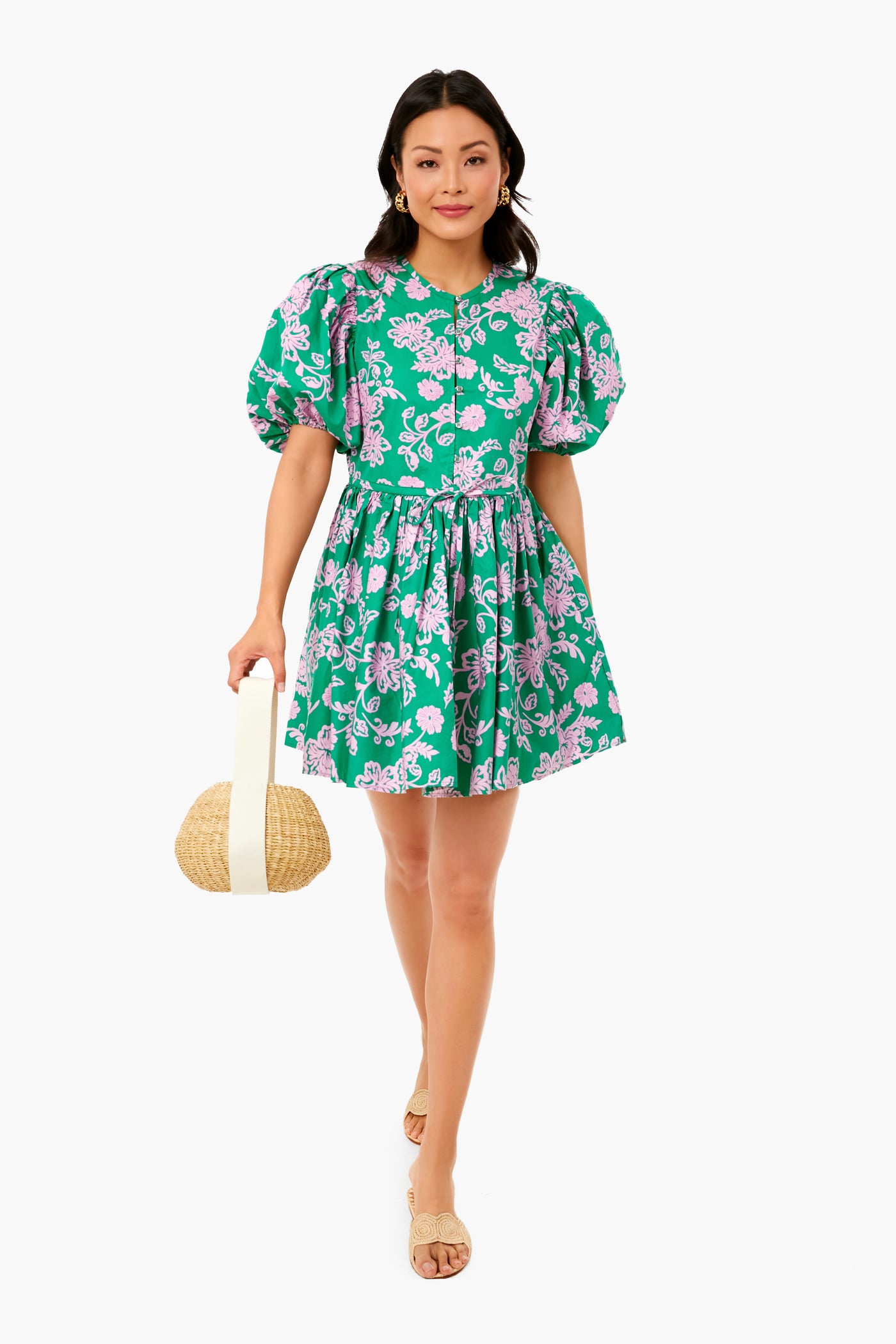 A woman in a XiRENA Caprisyn Green Aurie mini dress with pink floral pattern and puff sleeves, paired with beige sandals, holds a round straw handbag.