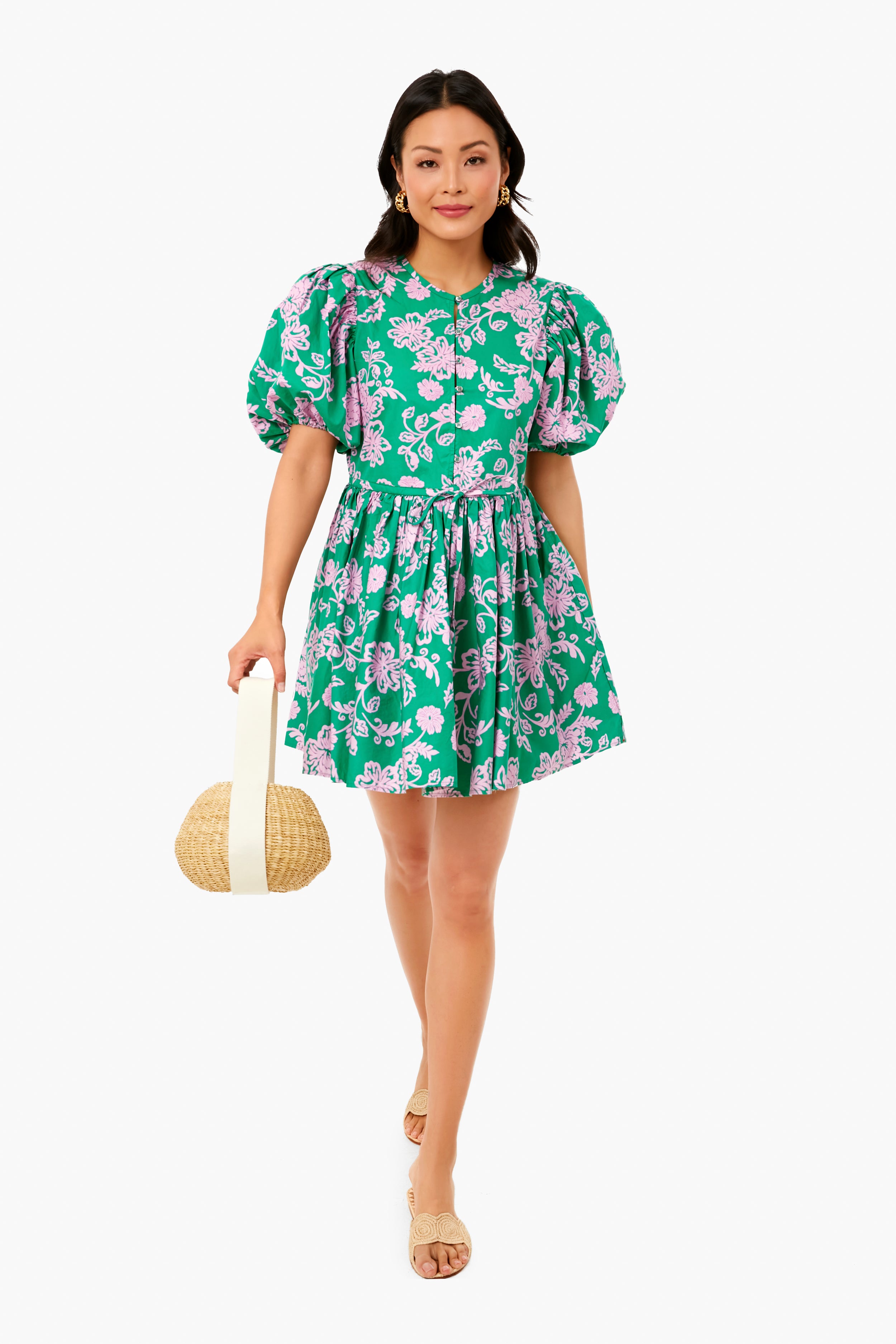 A woman in a XiRENA Caprisyn Green Aurie mini dress with pink floral pattern and puff sleeves, paired with beige sandals, holds a round straw handbag.